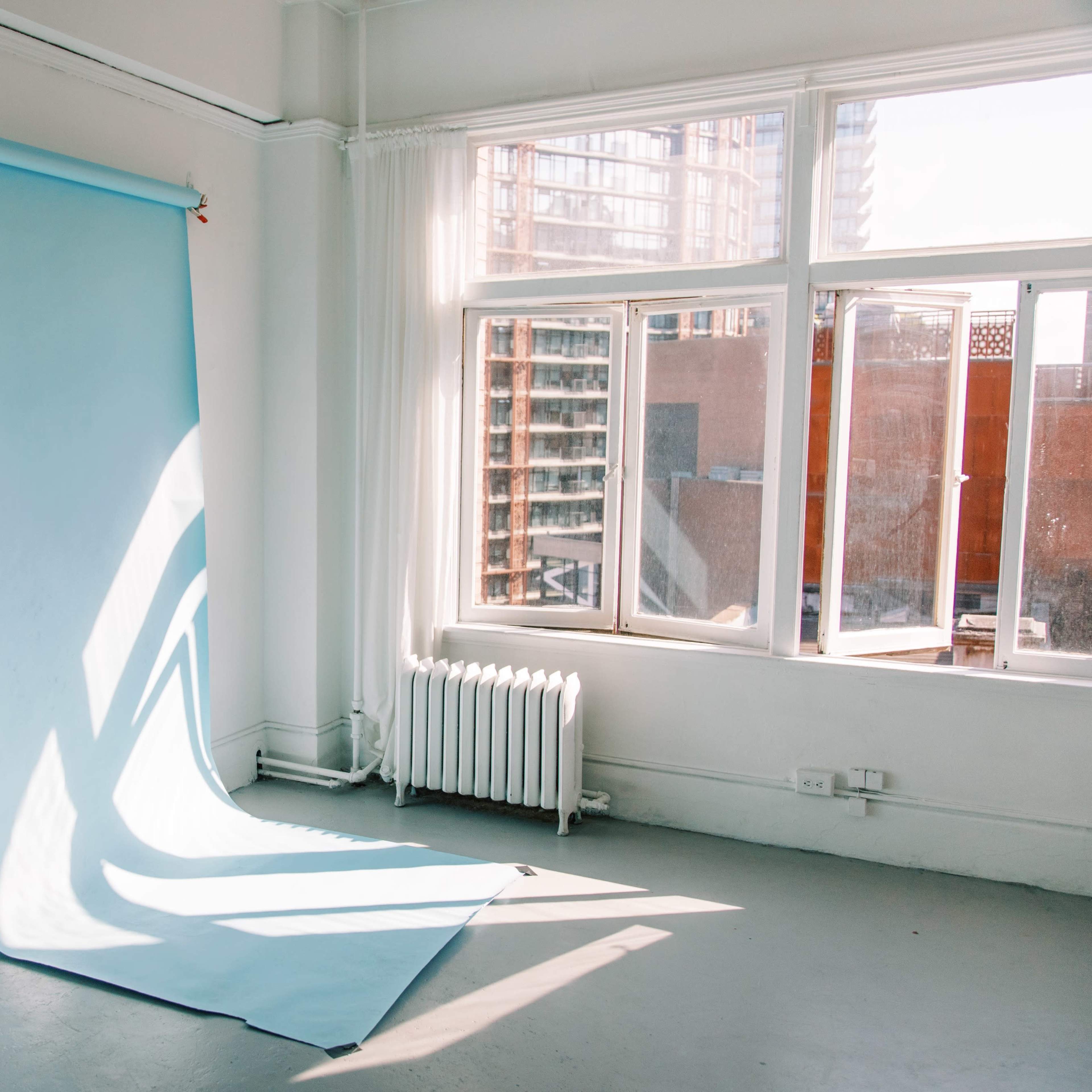 A light blue backdrop against a white wall in a sunlit room with large windows and a mirror.