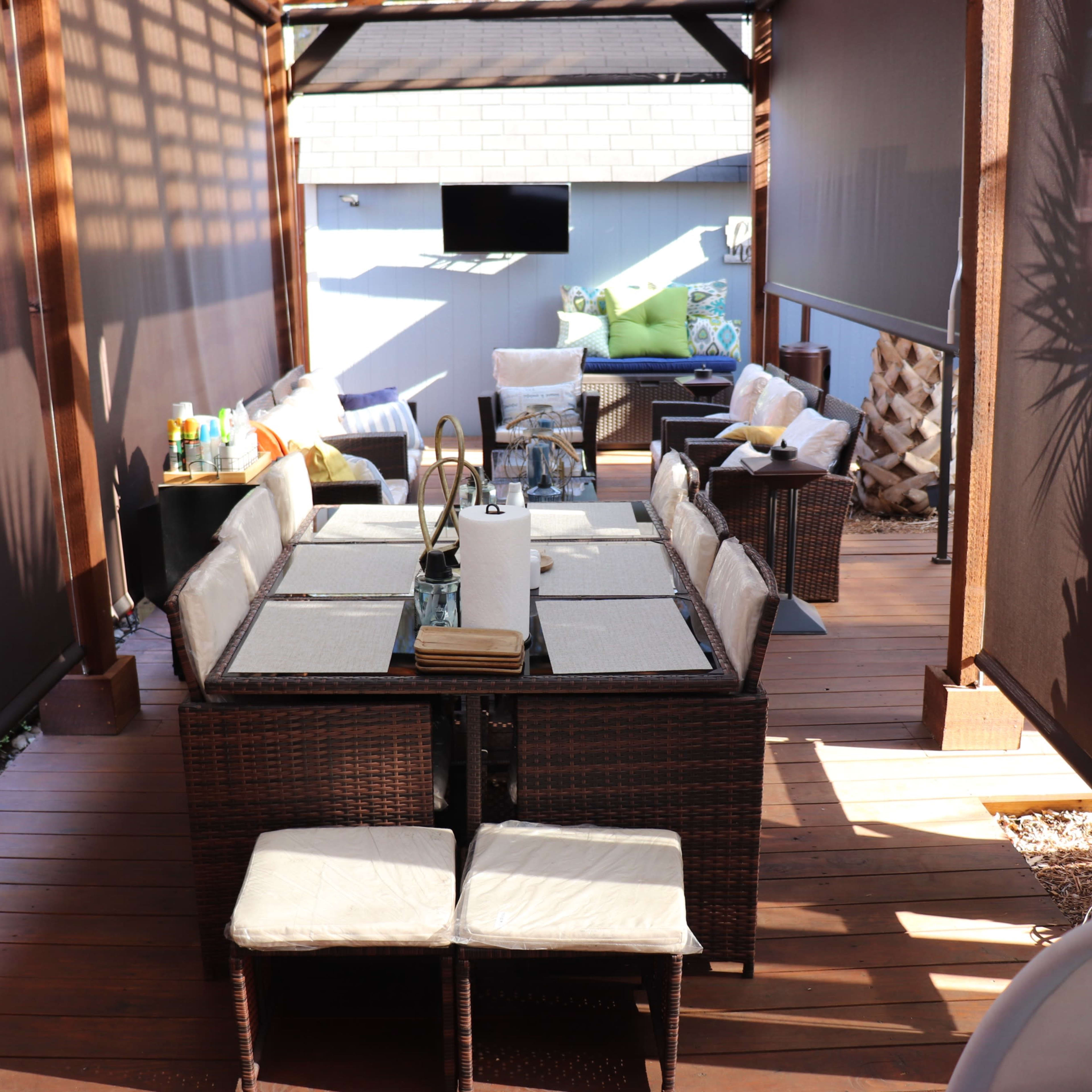 A shaded outdoor seating area featuring a large dining table surrounded by multiple chairs, with a small couch and decorative pillows in the background.
