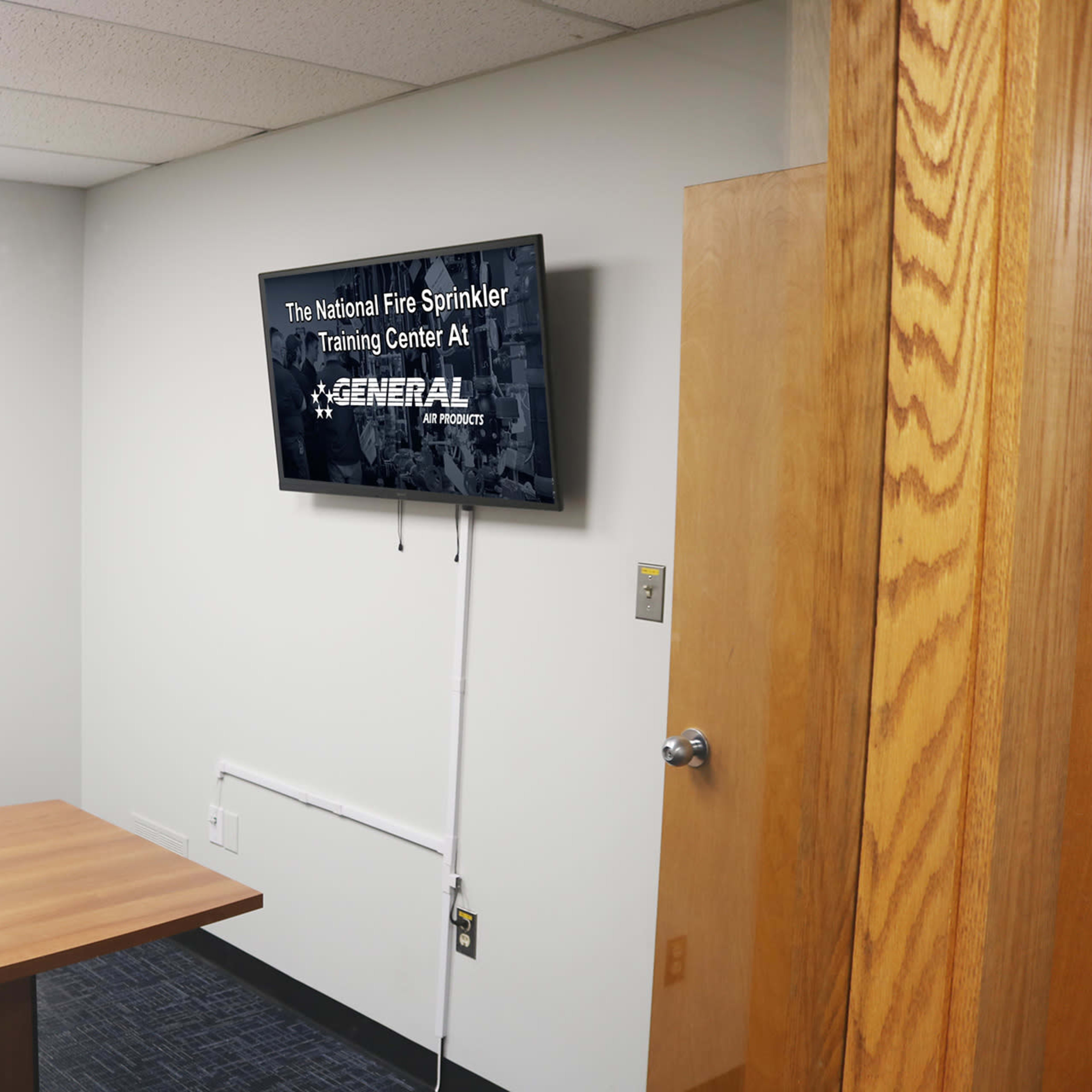 A training room with a wooden table, a chair, and a wall-mounted television displaying the title "The National Fire Sprinkler Training Center."