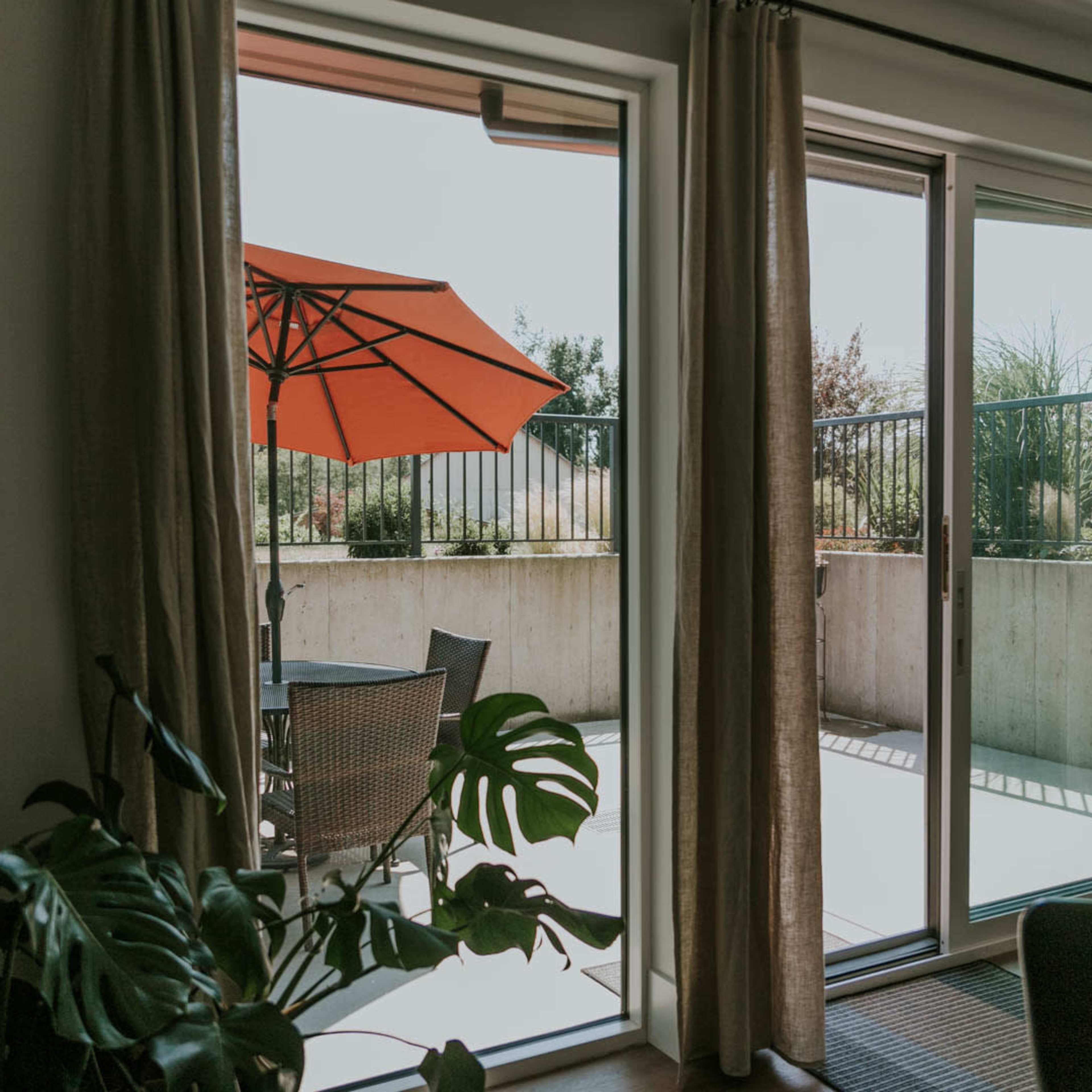 A bright living space features sliding glass doors leading to a patio with an orange umbrella over a small table and chairs.