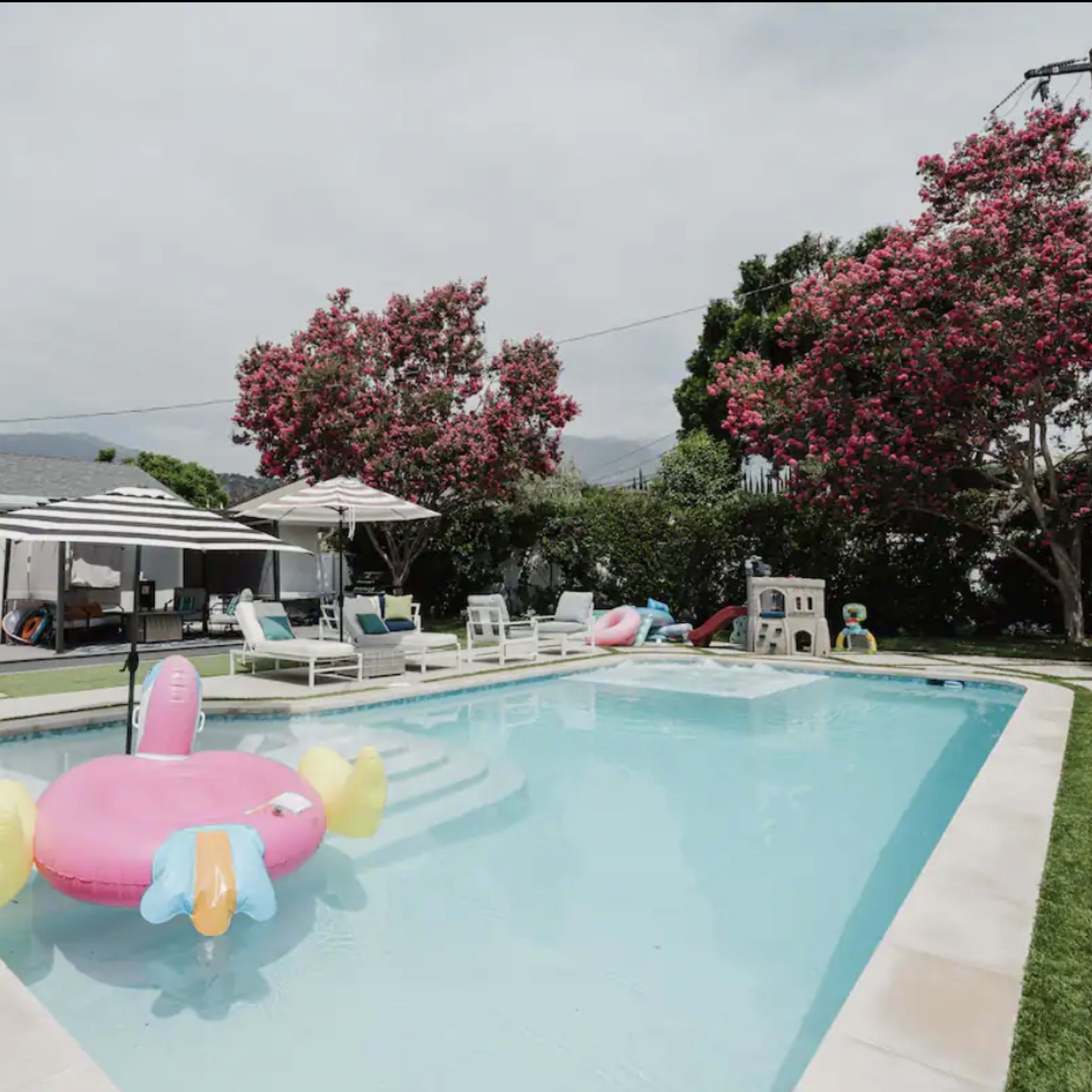 A swimming pool surrounded by lounge chairs and umbrellas, with colorful floaties resting on the water and blooming trees in the background.