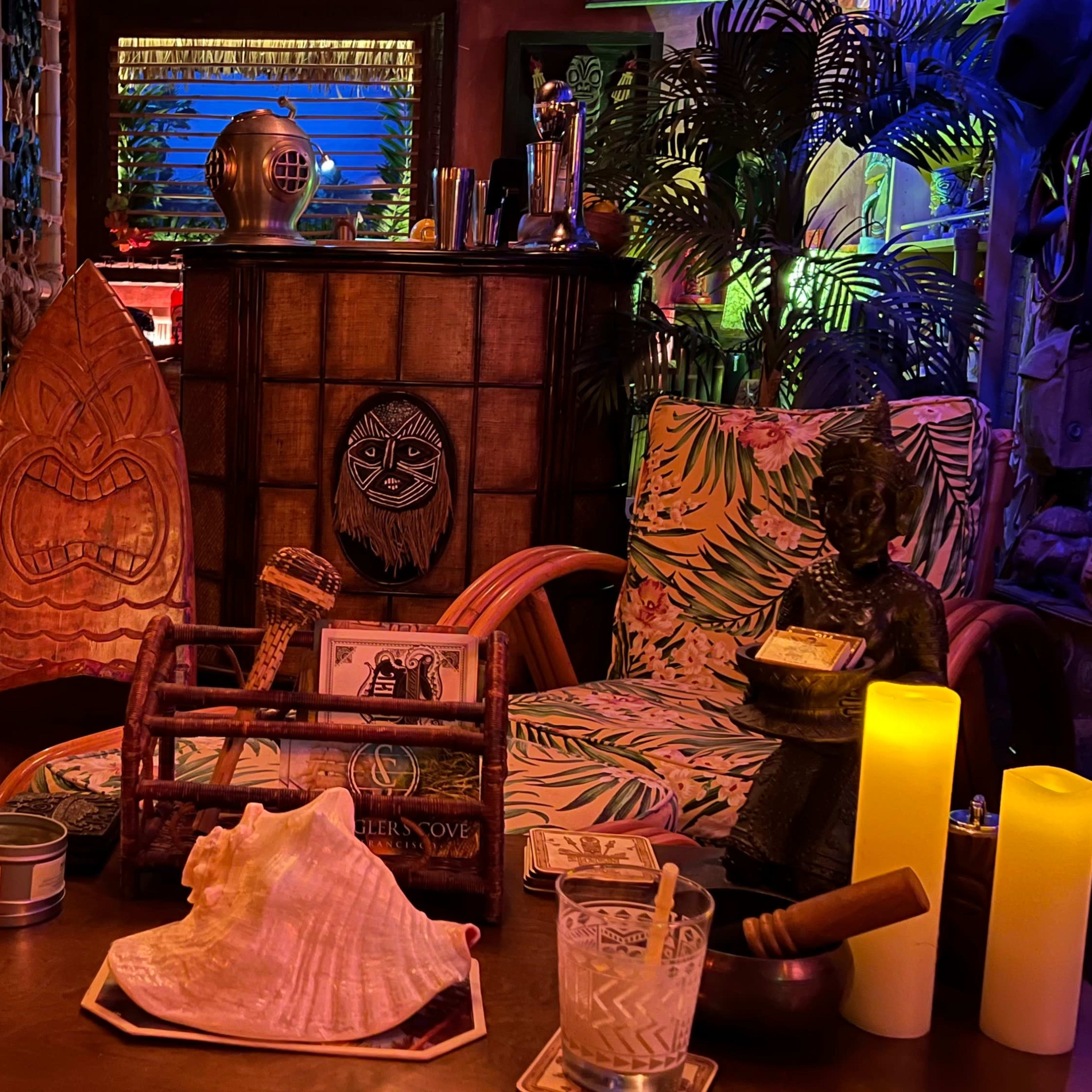 A dimly lit room decorated with tropical-themed furniture, a carved wooden figure, and various objects arranged on a wooden table.