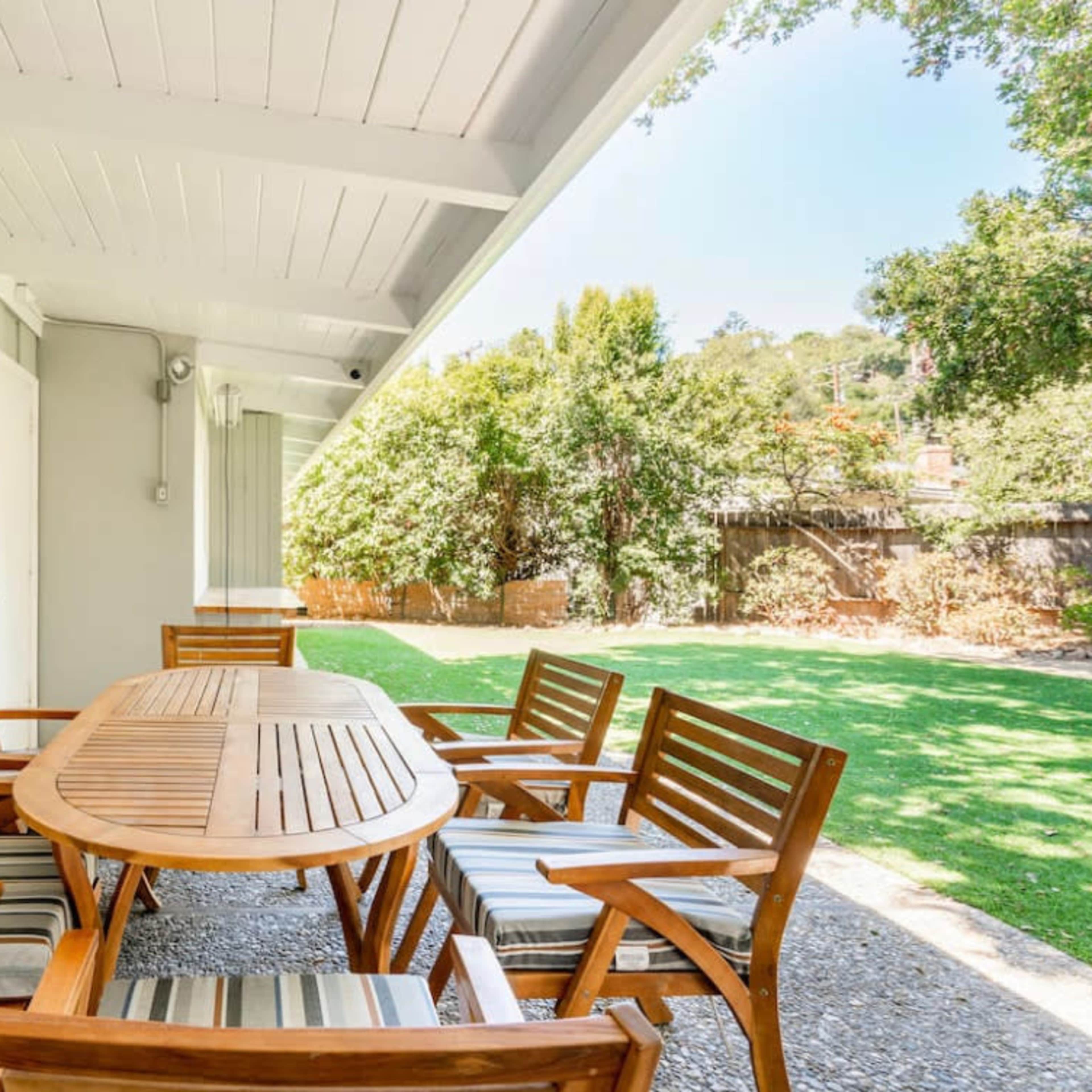 A wooden dining table surrounded by six chairs on a patio, overlooking a grassy yard and trees.