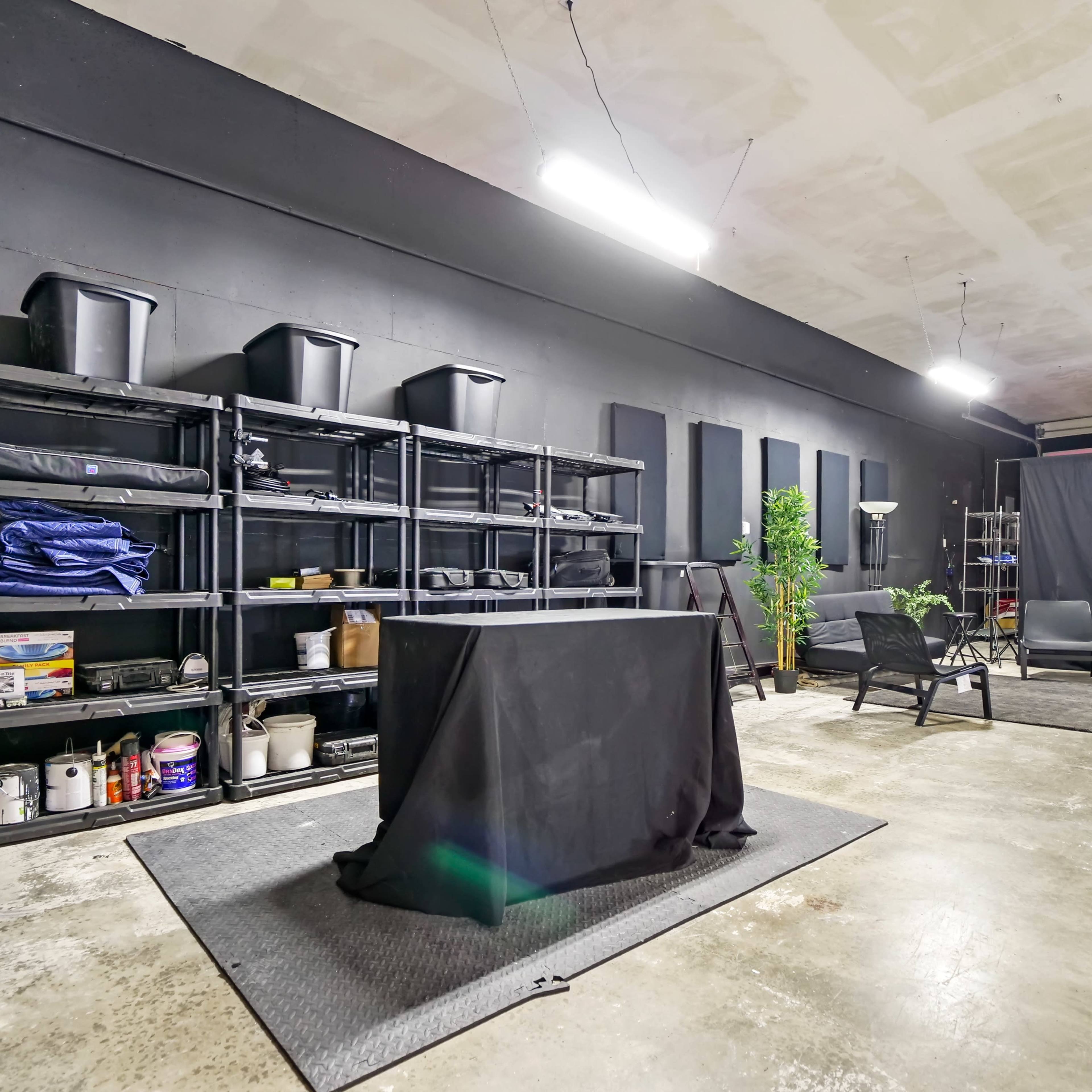 An industrial-style room with black walls, shelves filled with storage bins, a covered table in the center, and seating areas with minimal decor.