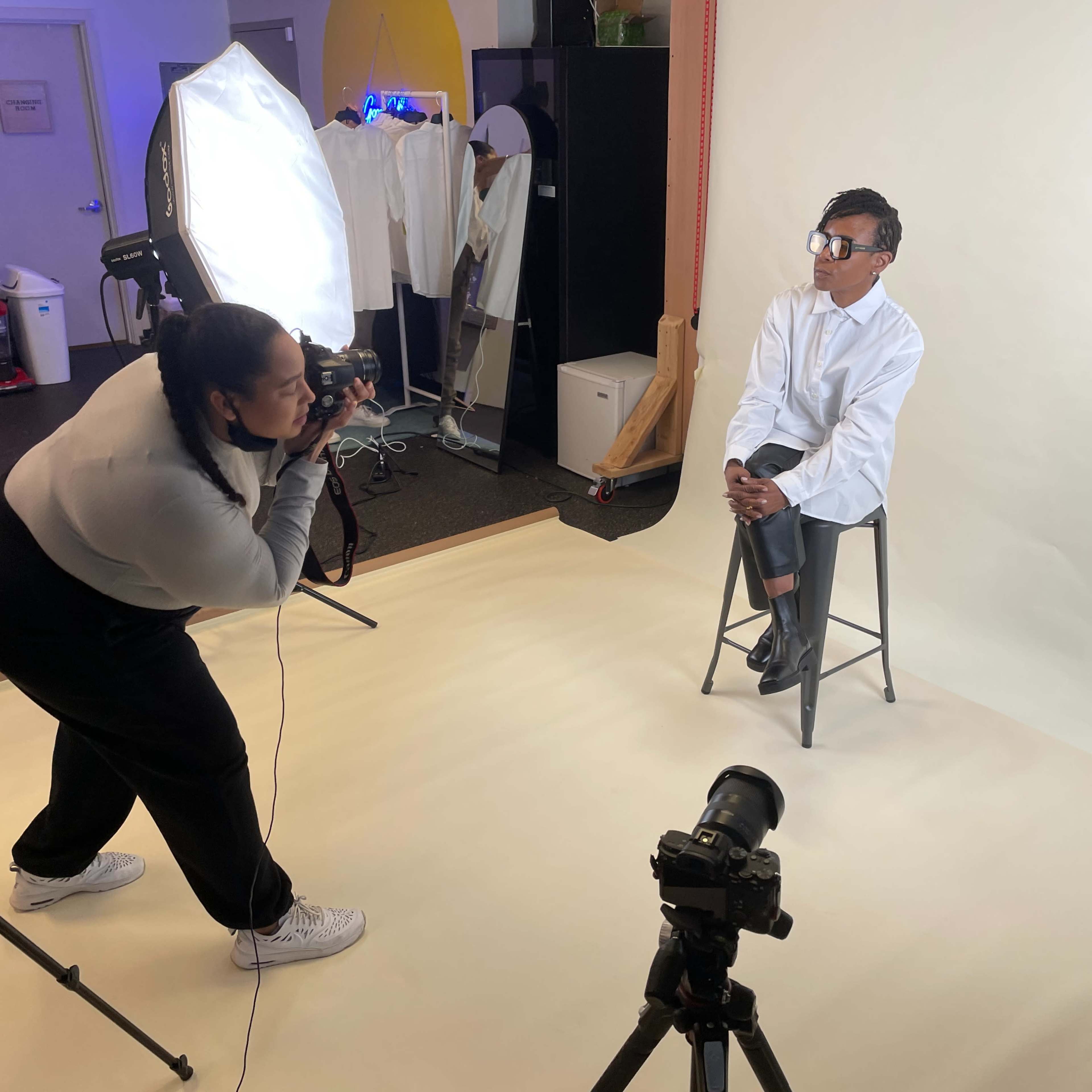 A photographer is taking a portrait of a model sitting on a stool in a studio with professional lighting equipment set up.