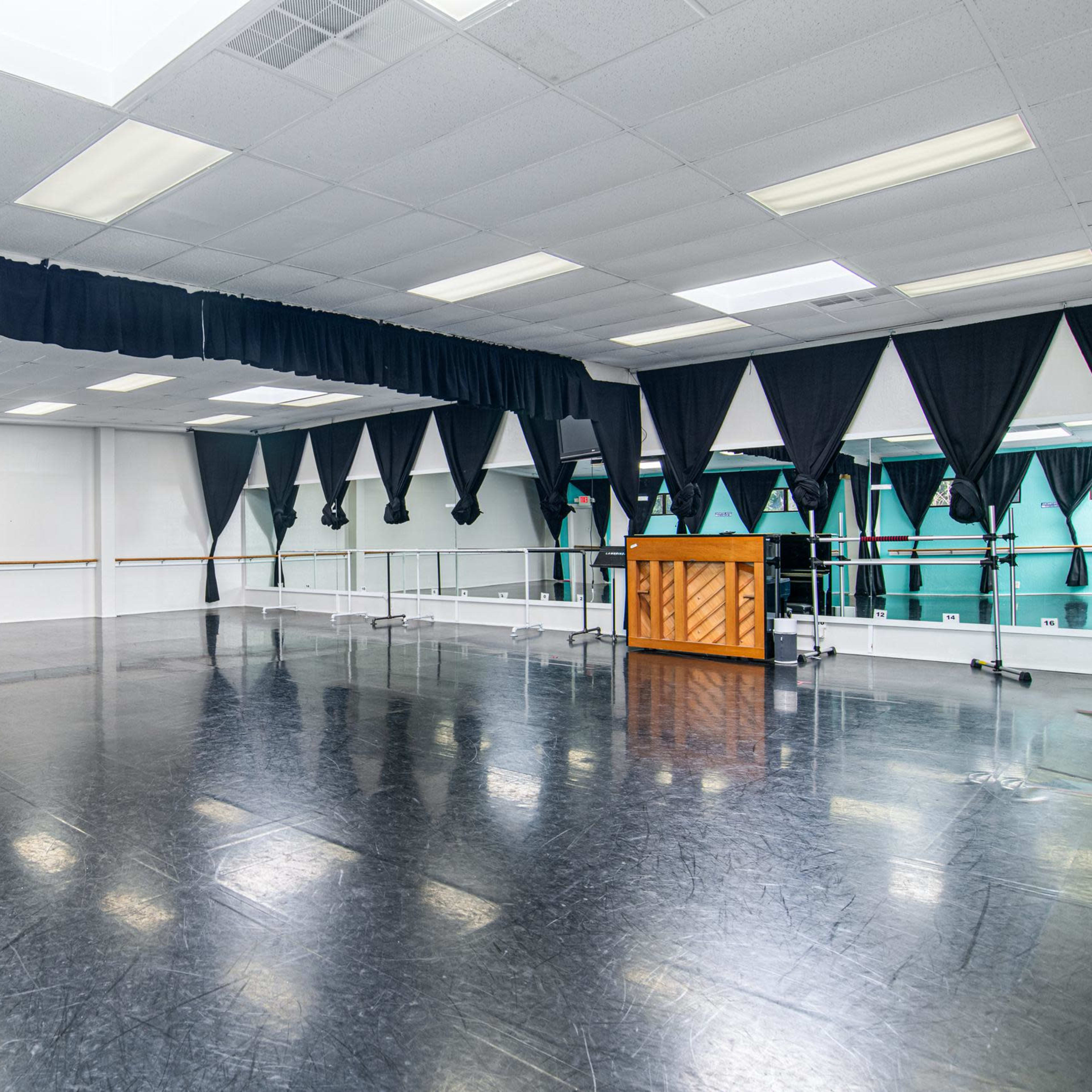 A spacious dance studio with dark flooring, large mirrors, and draped curtains, alongside a wooden piano positioned in the center.
