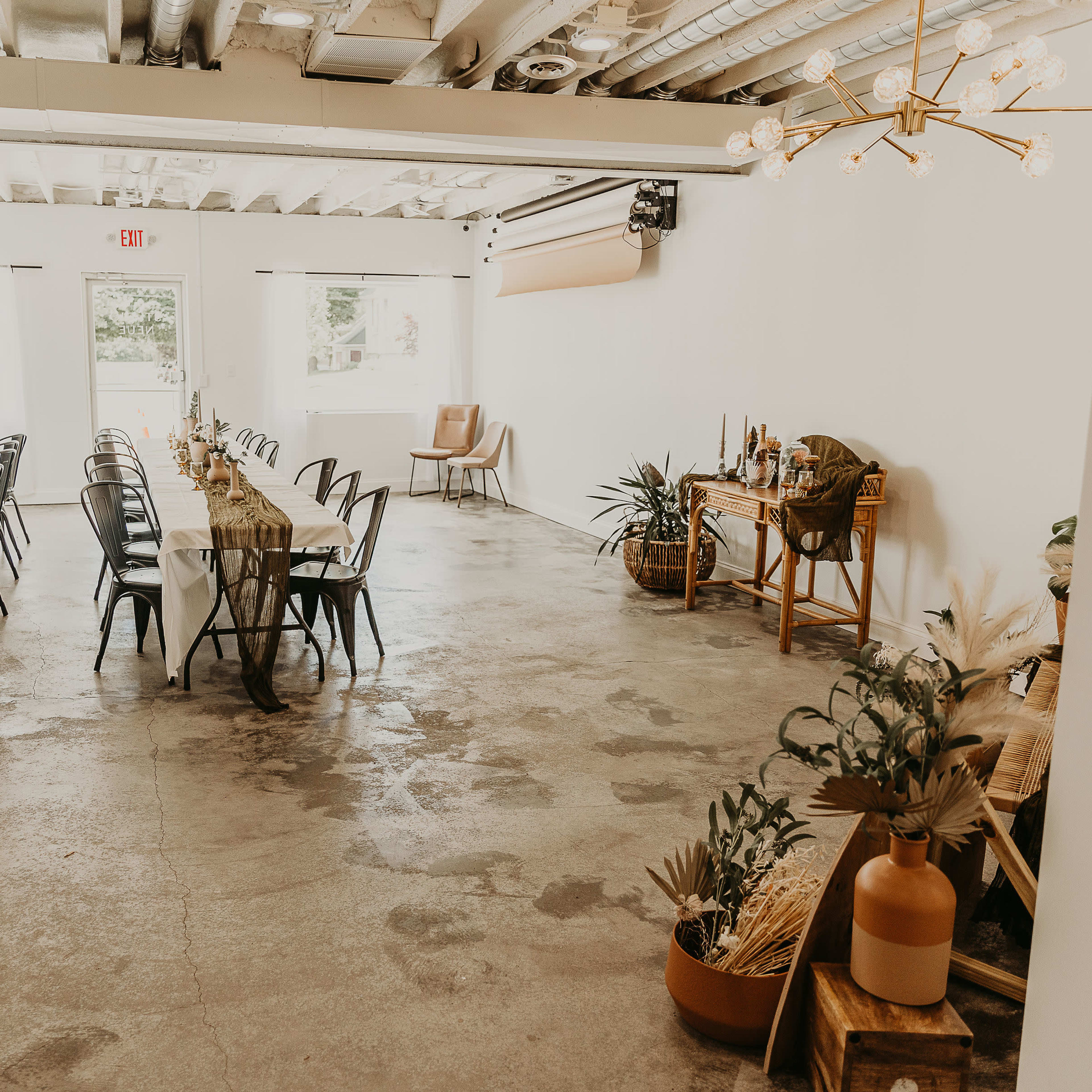 A spacious event room features long tables set with chairs, decorated with natural elements, and a refreshment area with plants.
