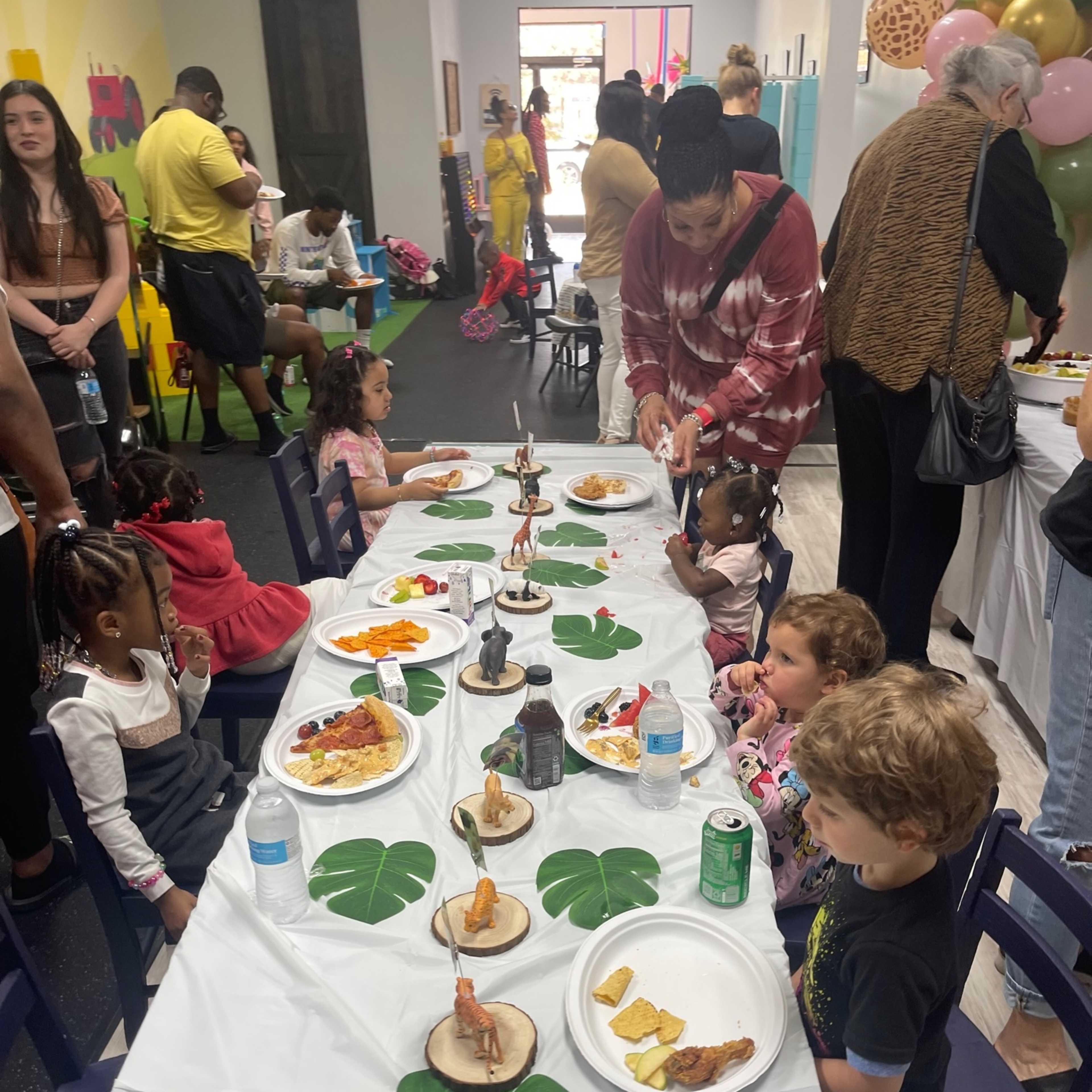 A long table is set for a children's party, featuring food and drinks, while kids sit and enjoy the meal.