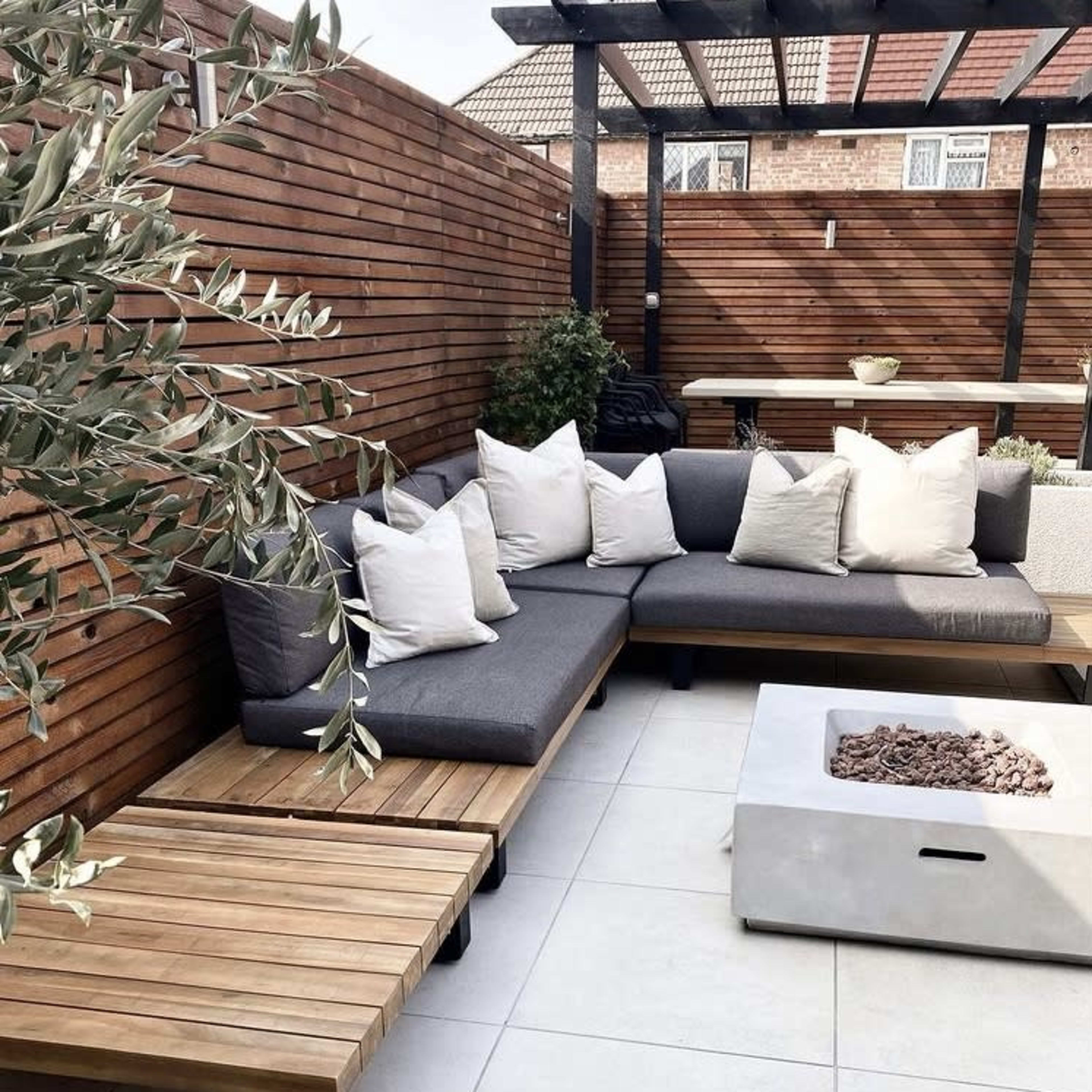 A outdoor seating area features a gray sectional sofa with white pillows, a wooden coffee table, and a modern fire pit surrounded by gray tiles and wooden fencing.
