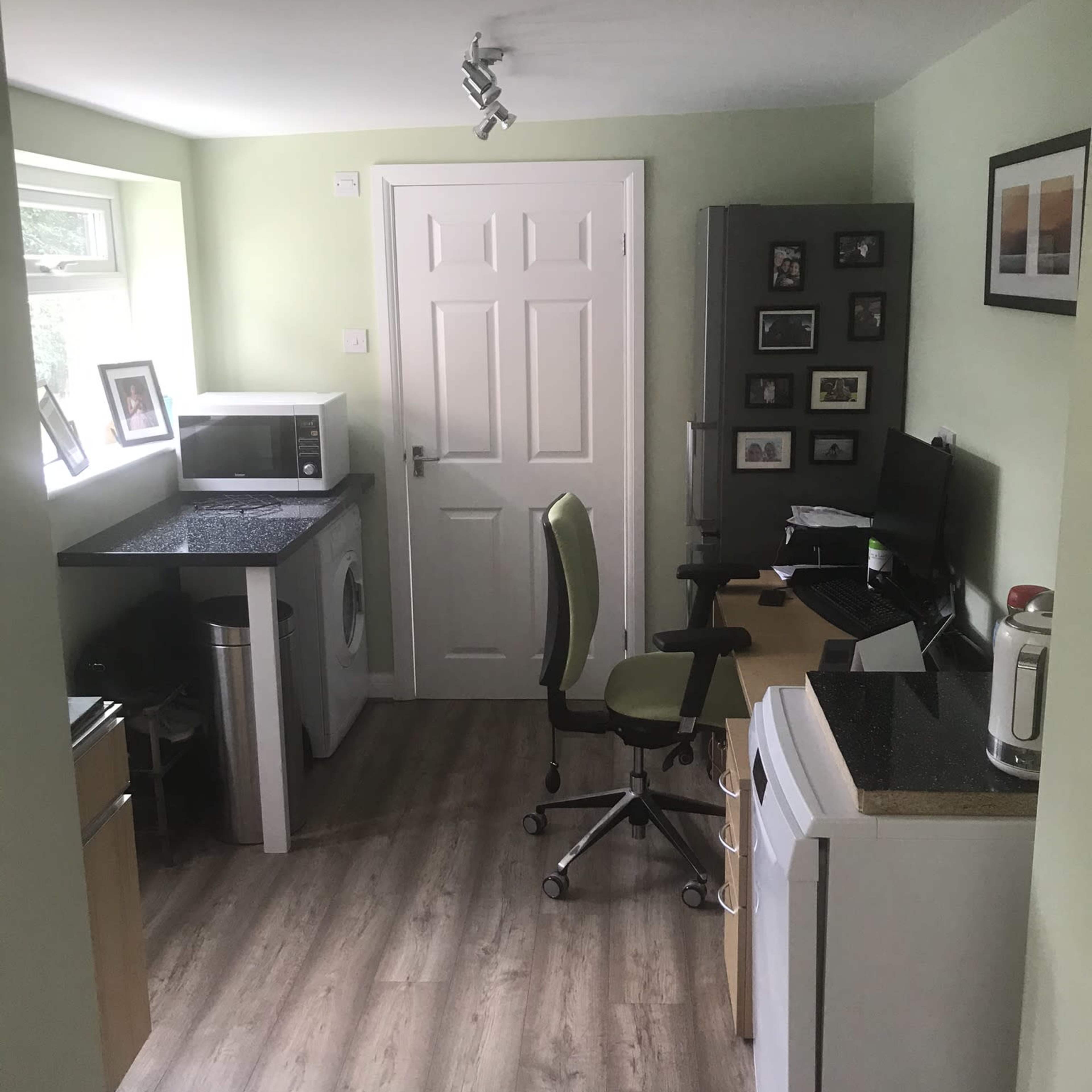 A small, functional kitchen area with a washing machine, microwave, refrigerator, and a desk with a computer and office chair.