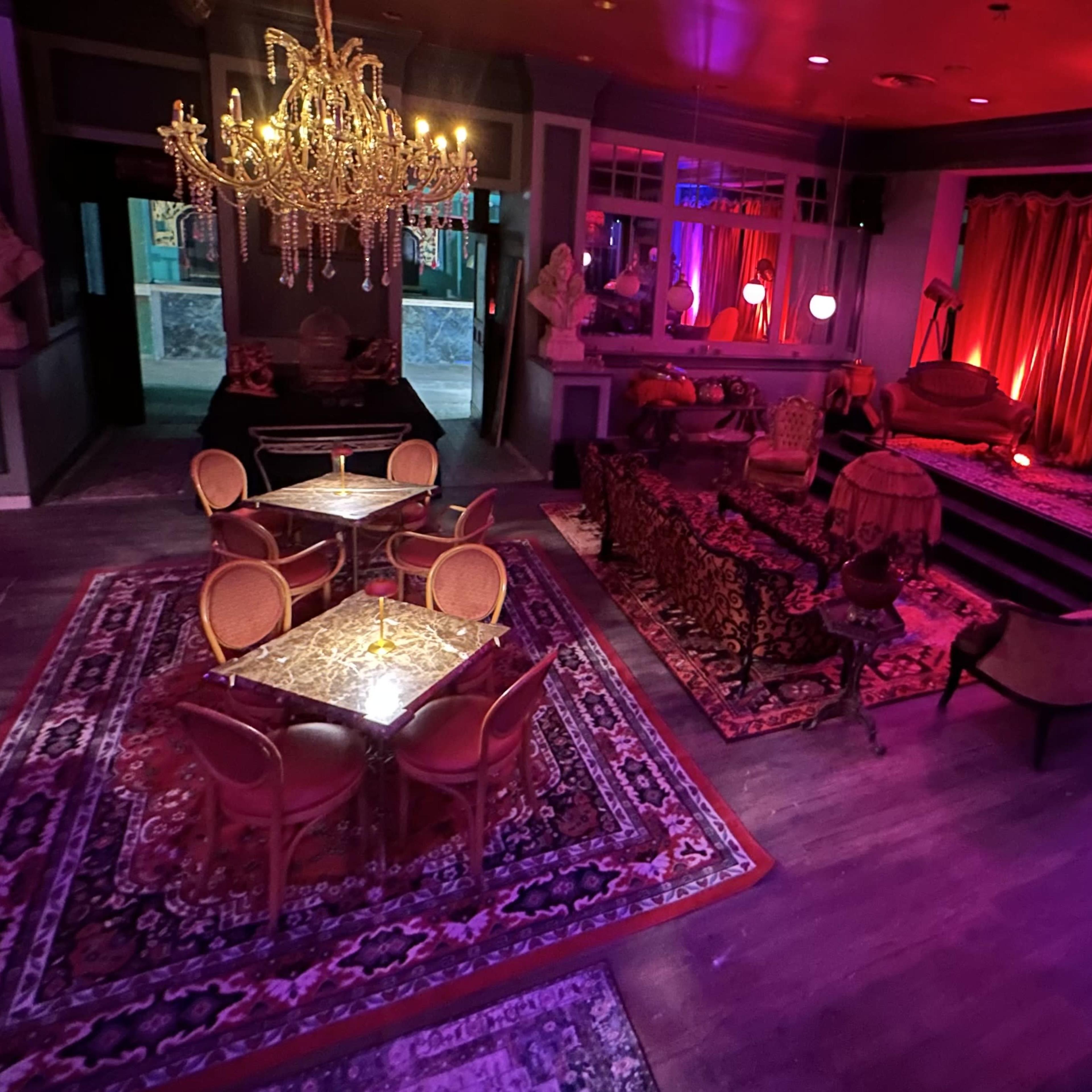 A dimly lit lounge with ornate furniture, a chandelier, and colorful lighting, featuring tables and chairs arranged on a patterned rug.