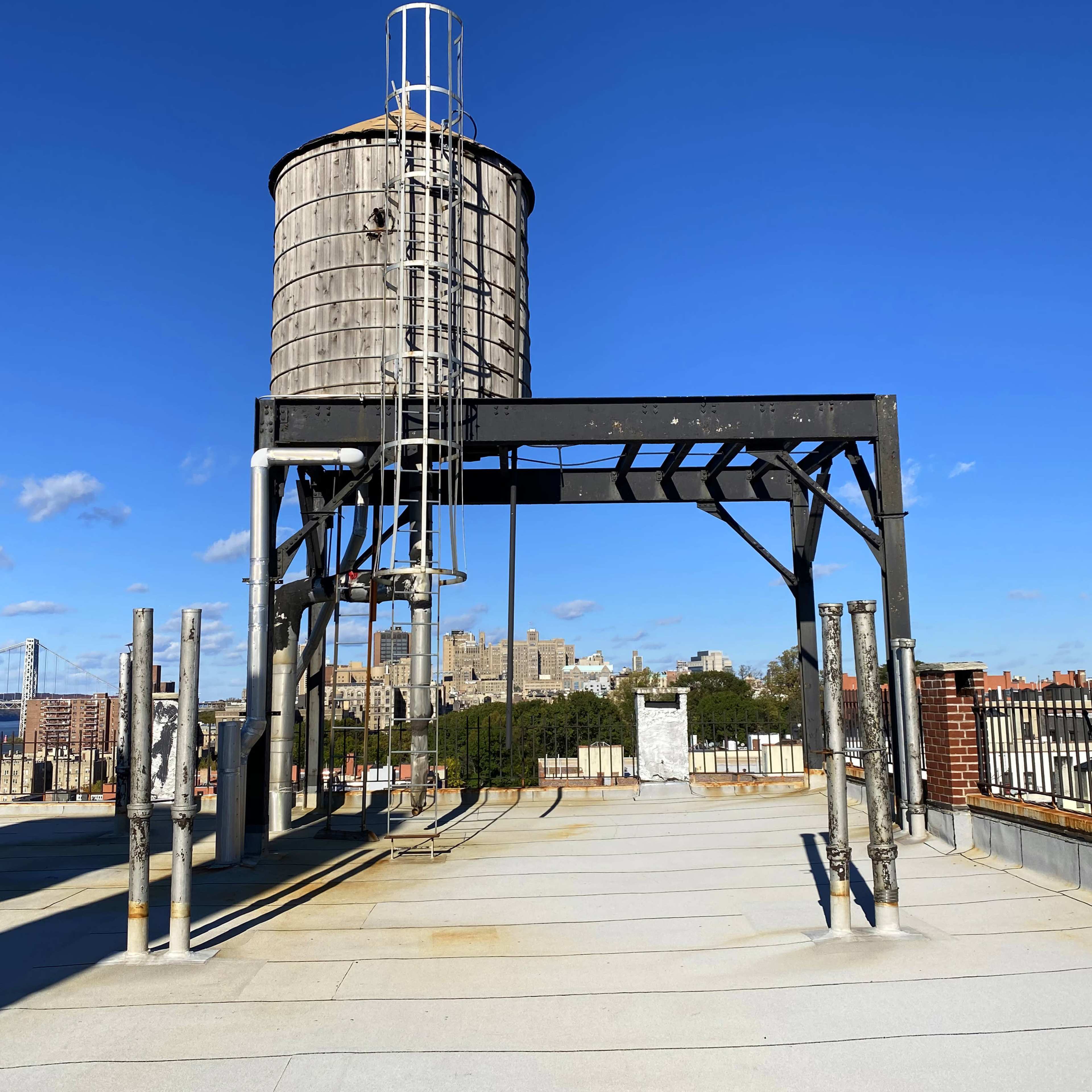 A wooden water tower stands on a rooftop with a clear blue sky and a city skyline in the background.