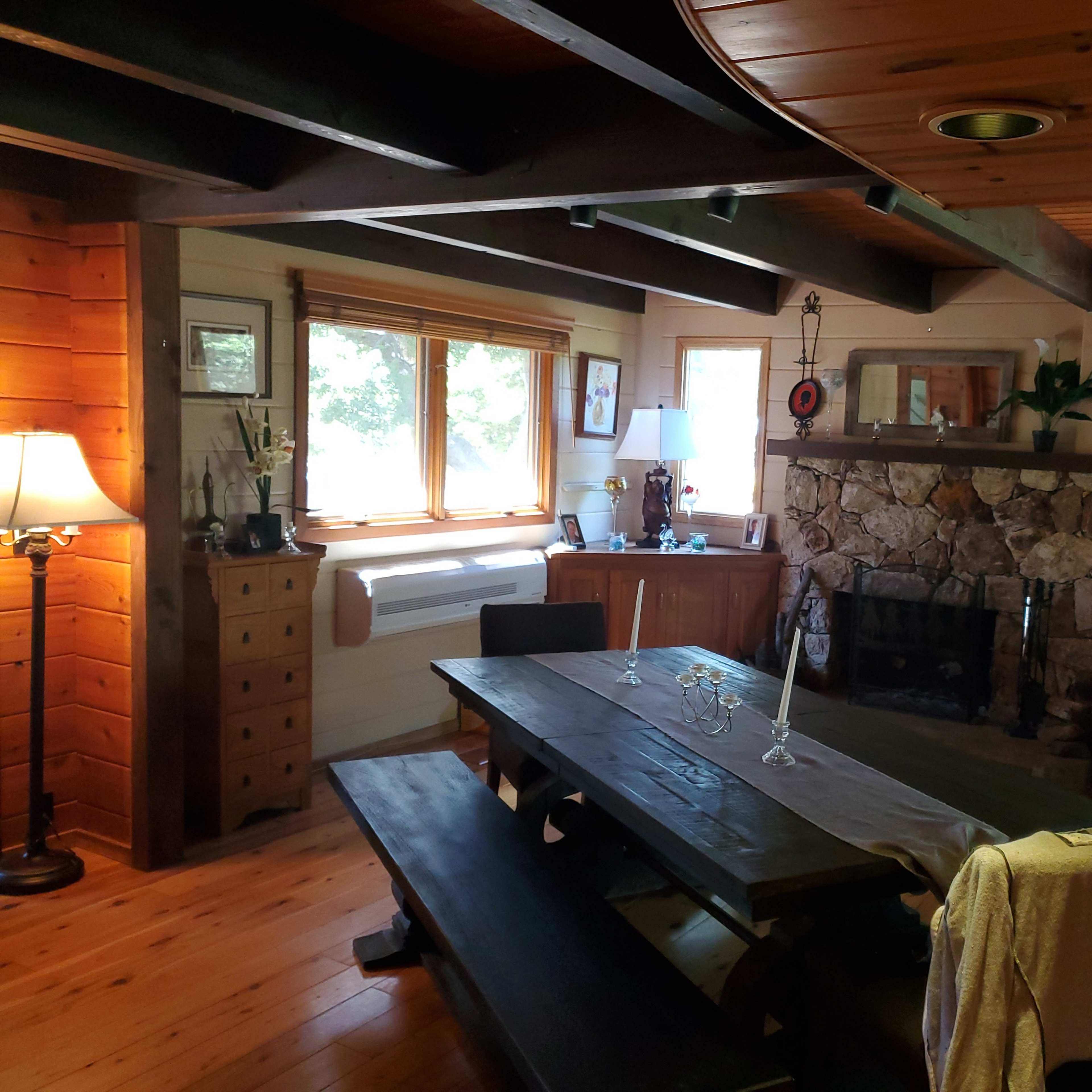 A wooden dining area with a large table and stone fireplace, illuminated by natural light from the windows.