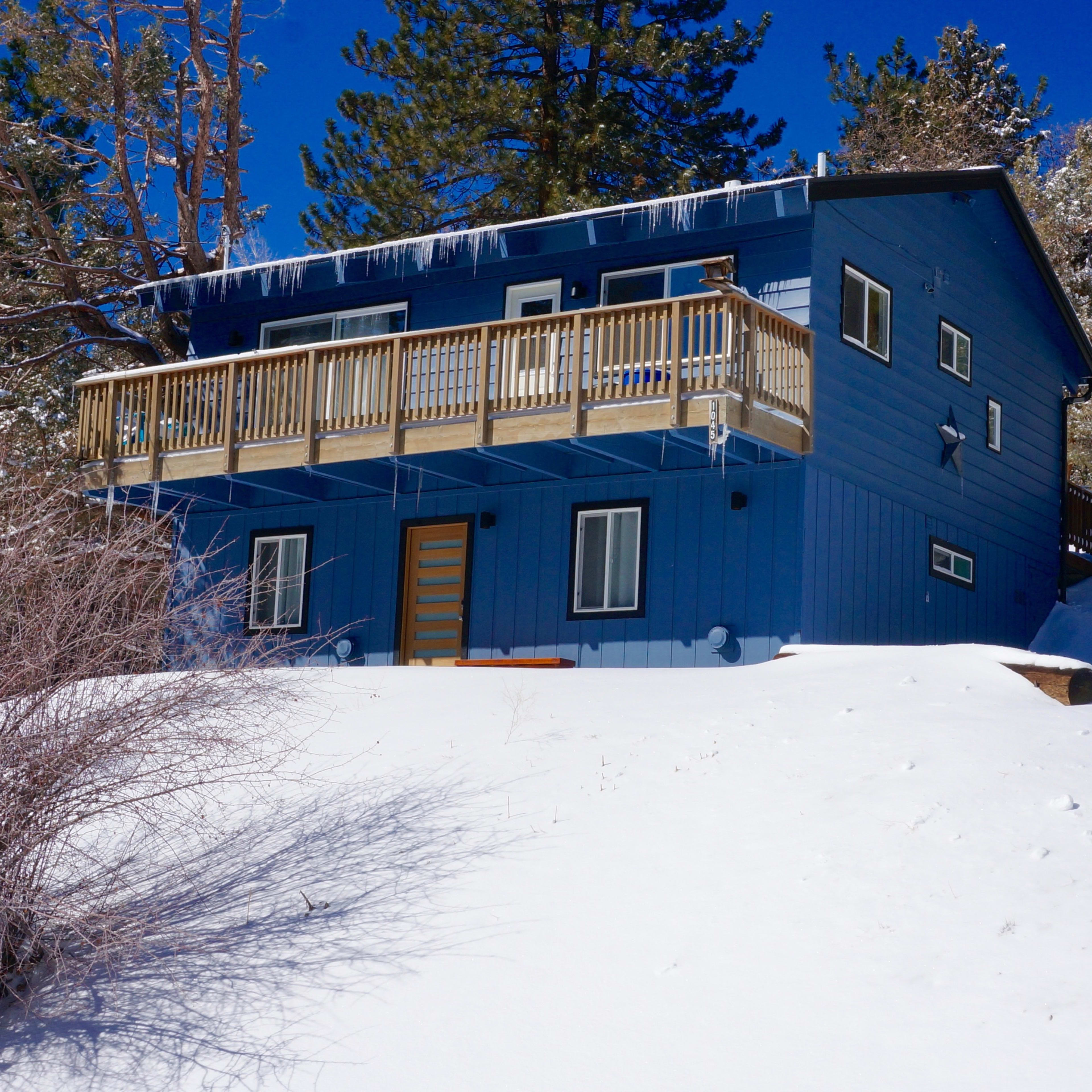 A blue two-story house sits on a snowy hillside, surrounded by trees and a sign at the front.