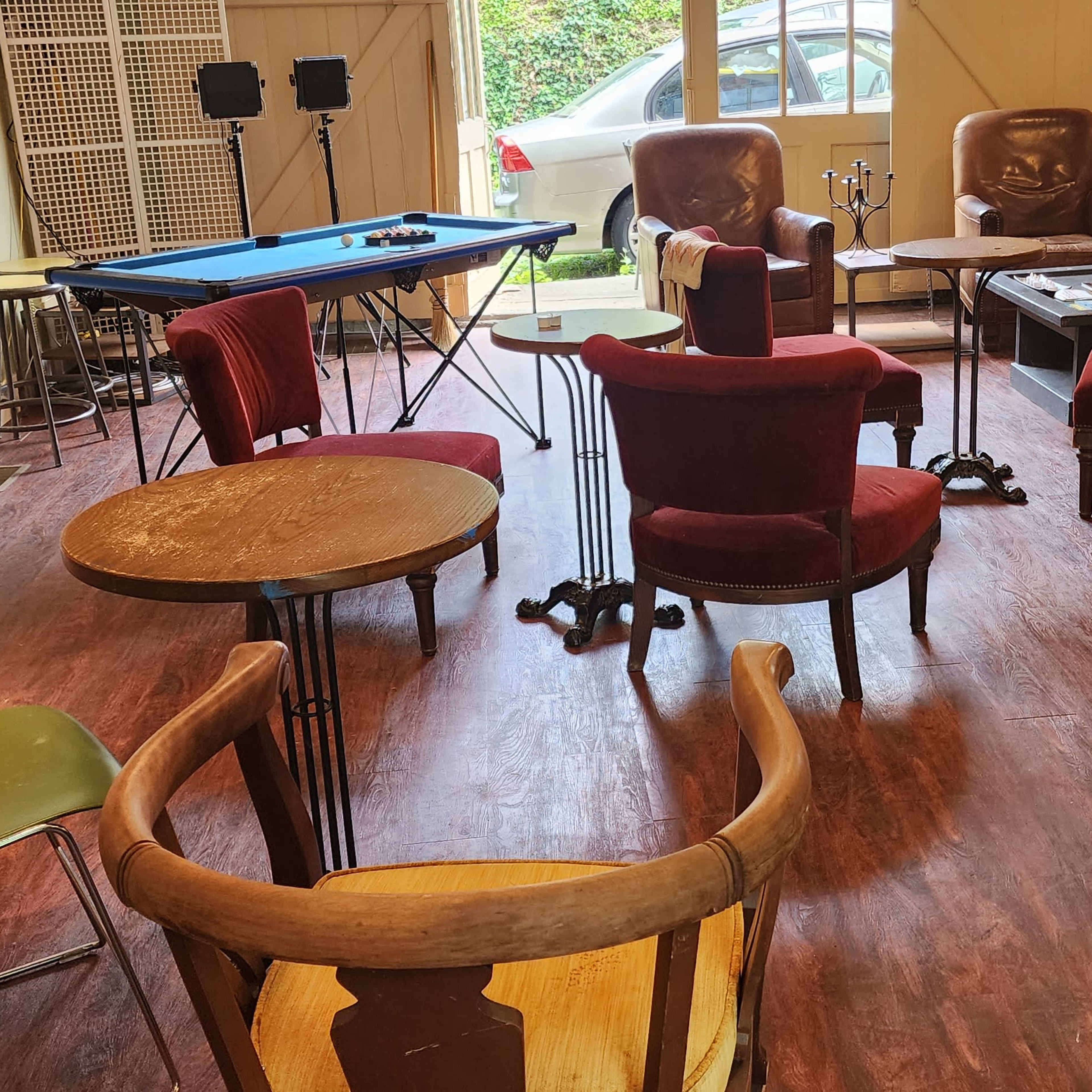The scene shows a spacious room with several chairs and tables arranged around a blue pool table, with a couch and a car visible outside through the windows.