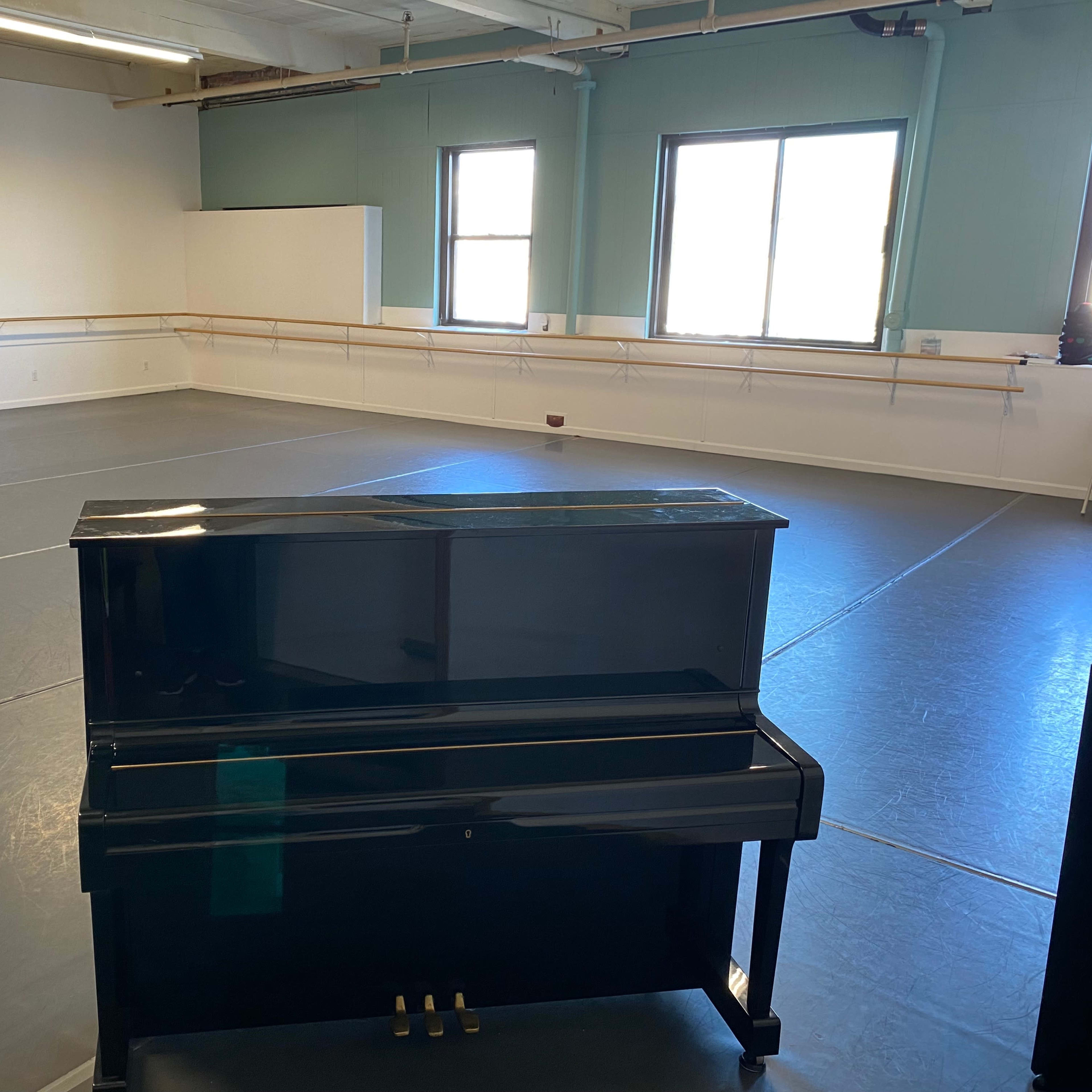 A black upright piano stands in an empty dance studio with gray flooring and large windows.