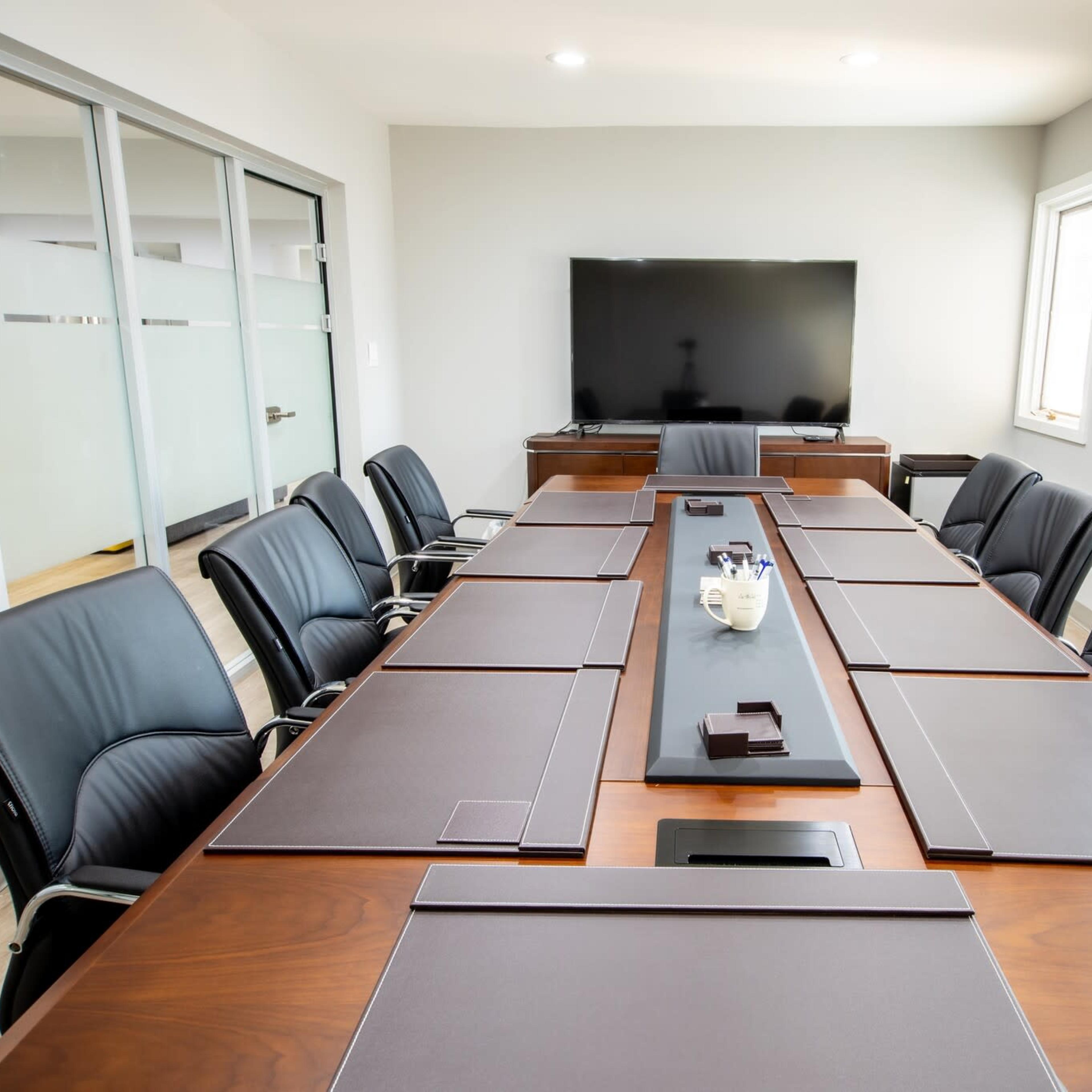 A modern conference room features a long wooden table surrounded by black leather chairs, with a large screen on the wall and natural light coming through windows.