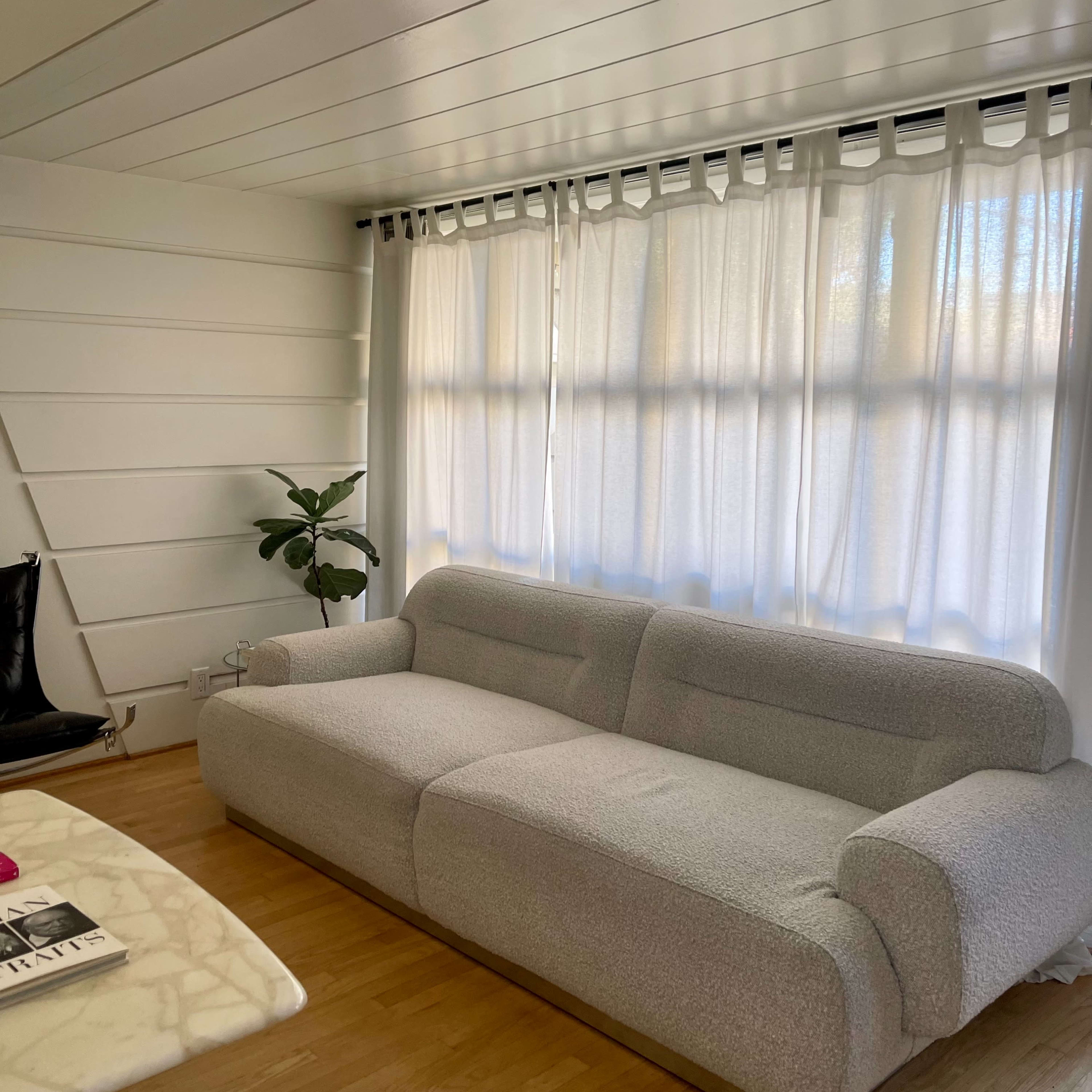 A spacious living room features a gray sofa, a marble coffee table, and large windows covered with white curtains.