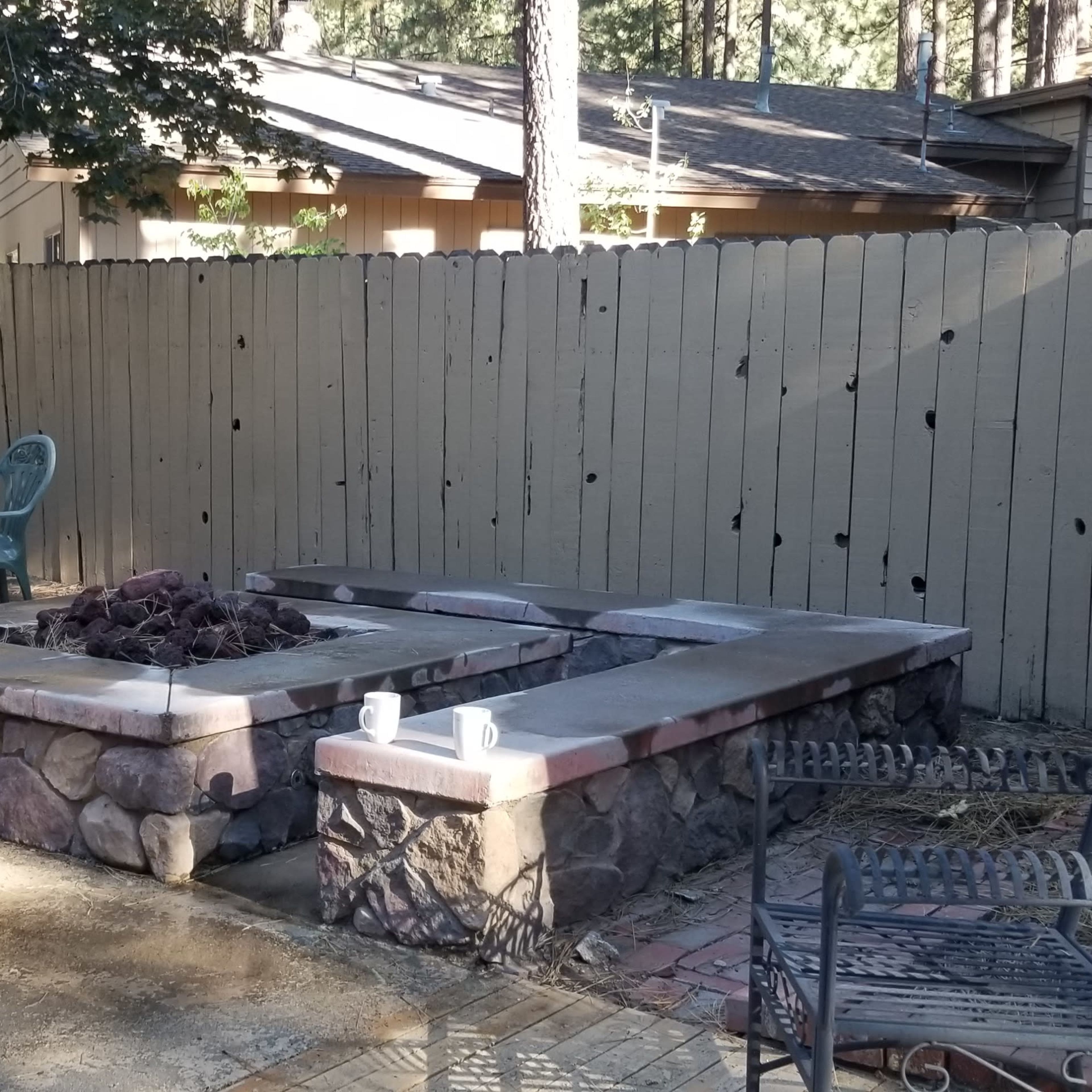 A backyard with a stone fire pit surrounded by a concrete patio, a grill, and several chairs against a wooden fence.