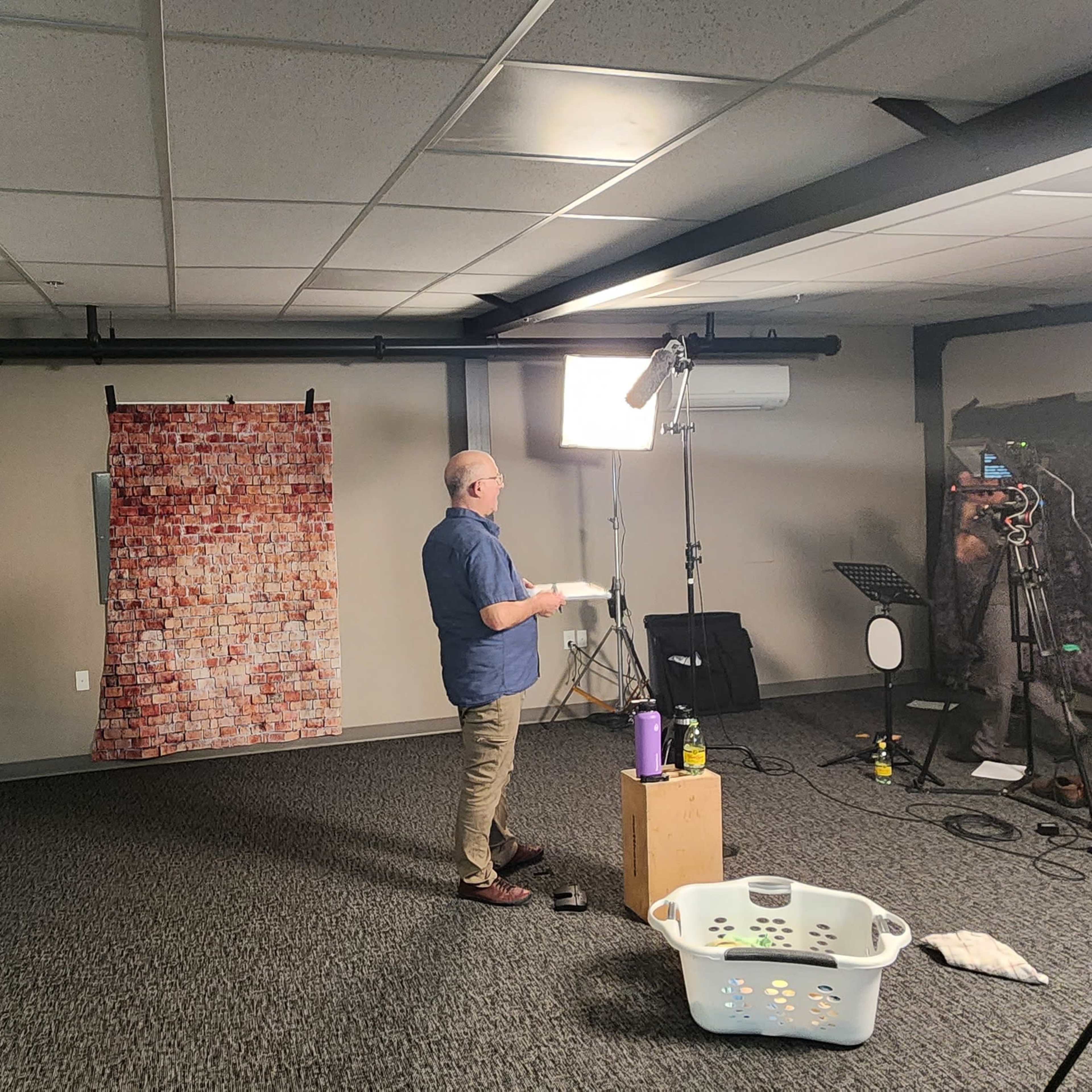 A man stands in a studio setting with various lighting equipment and backdrops while holding a clipboard.