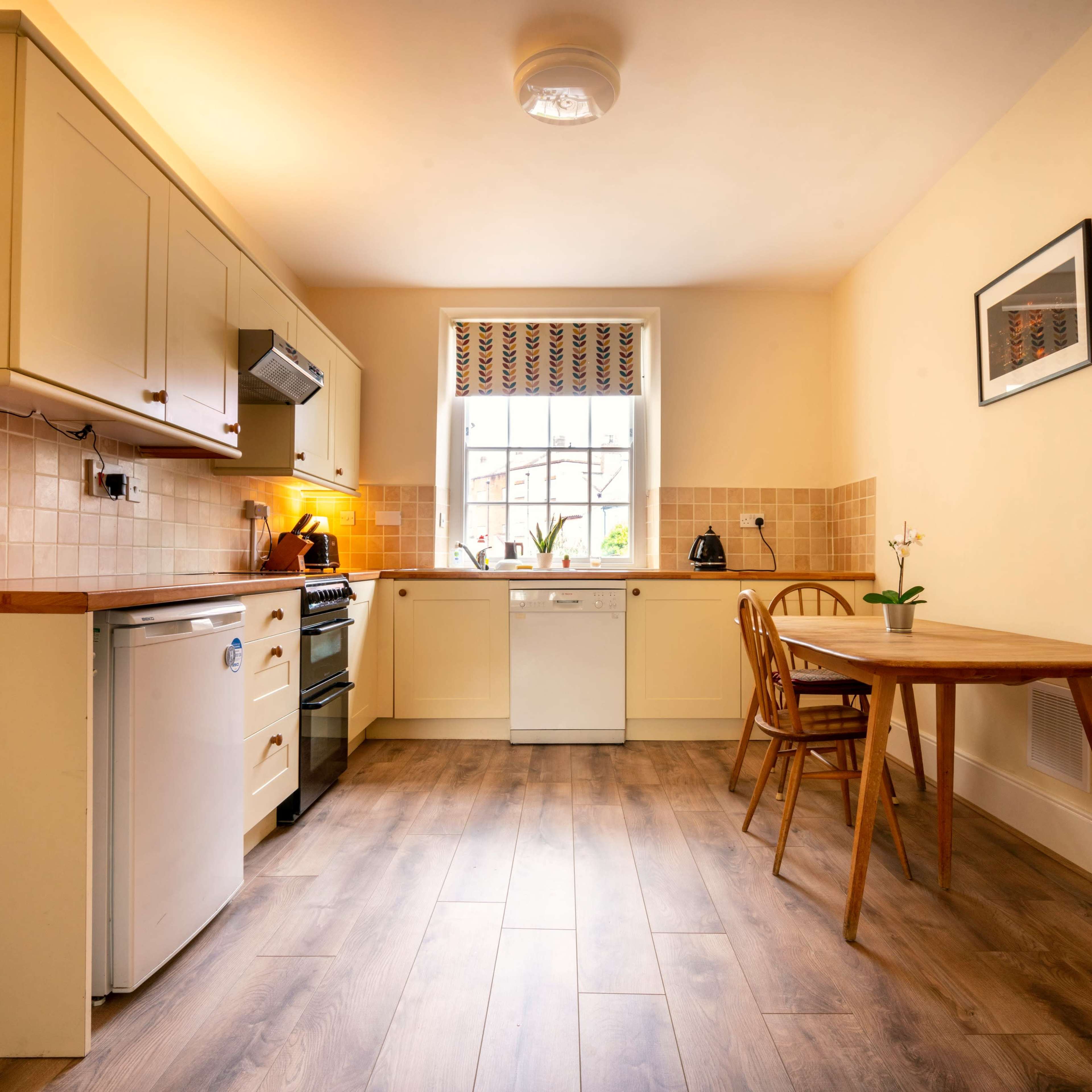 A bright kitchen features beige cabinetry, a window with checked curtains, and a wooden dining table with a chair.