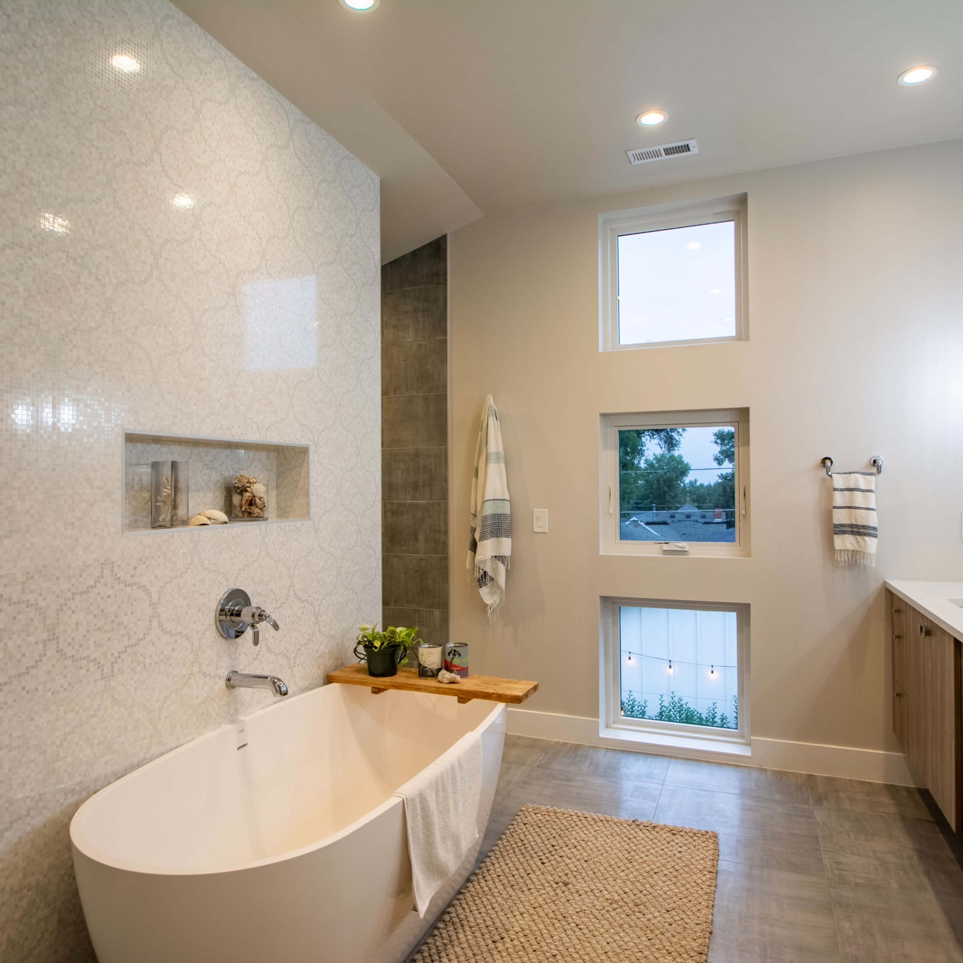 A modern bathroom featuring a freestanding bathtub, a geometric tiled accent wall, ample natural light from small windows, and a dual sink vanity with wooden cabinetry.