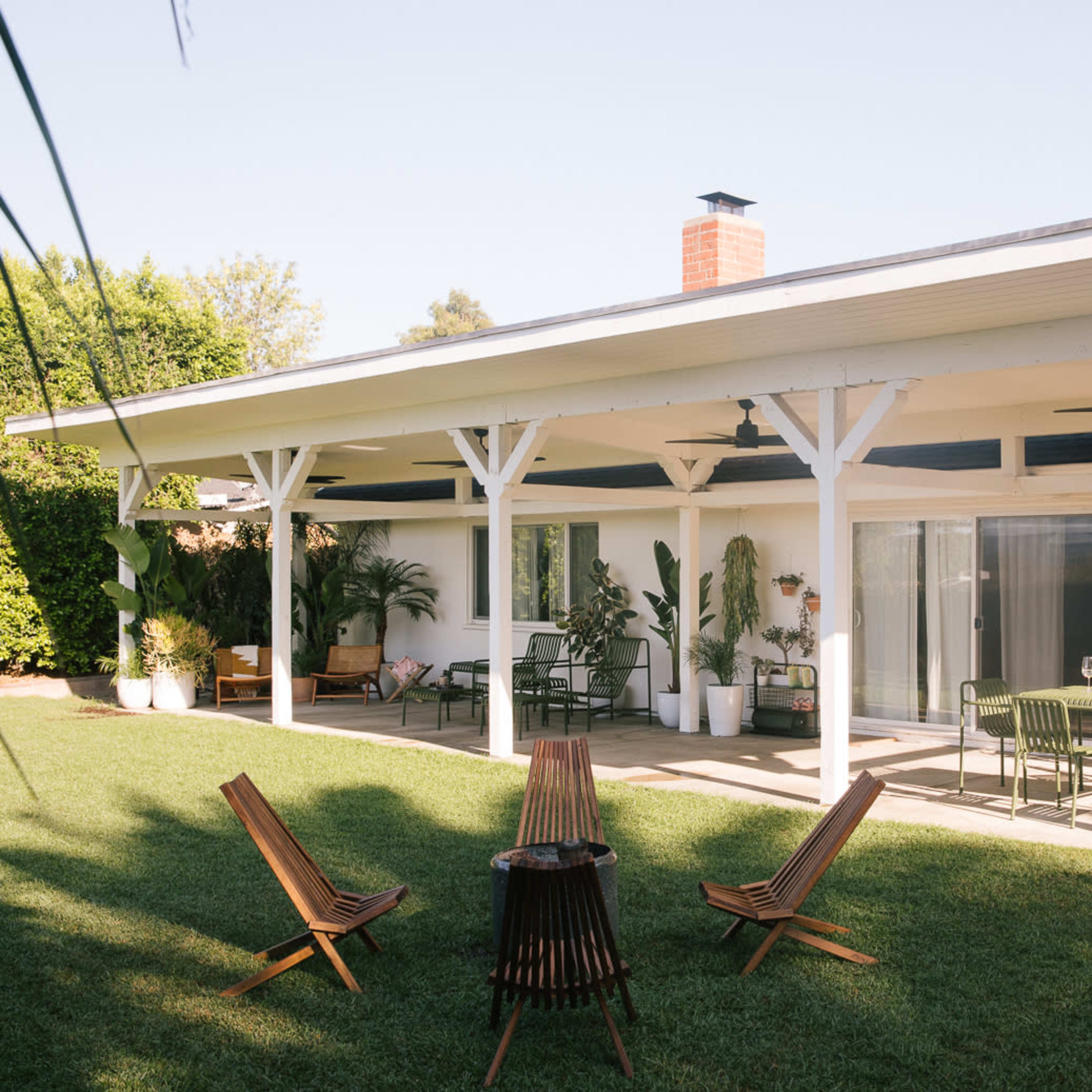 A spacious backyard features a covered patio with seating and a dining area surrounded by potted plants and a well-maintained lawn.