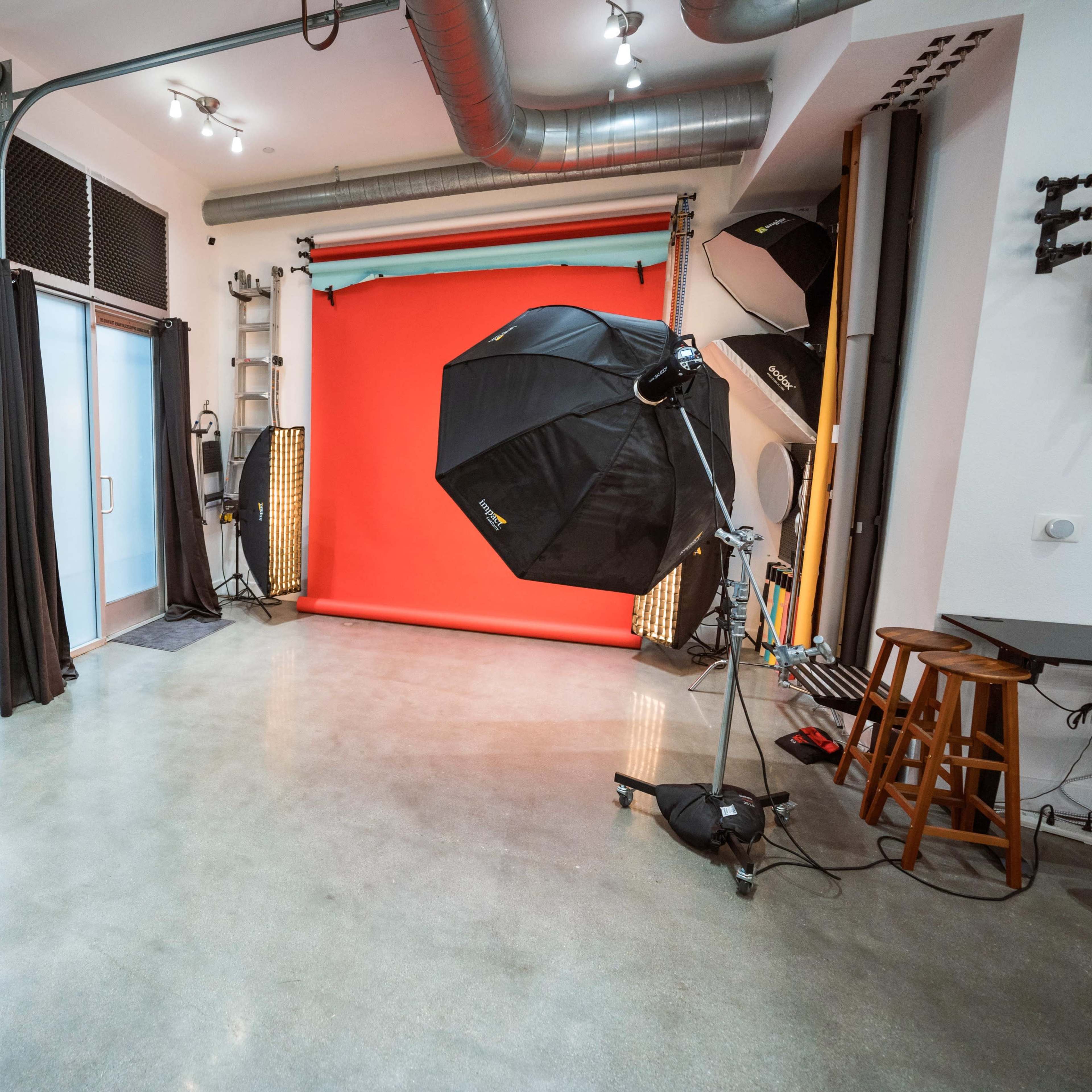 A photography studio with a bright red backdrop, an umbrella light setup, and various equipment arranged in a modern space.