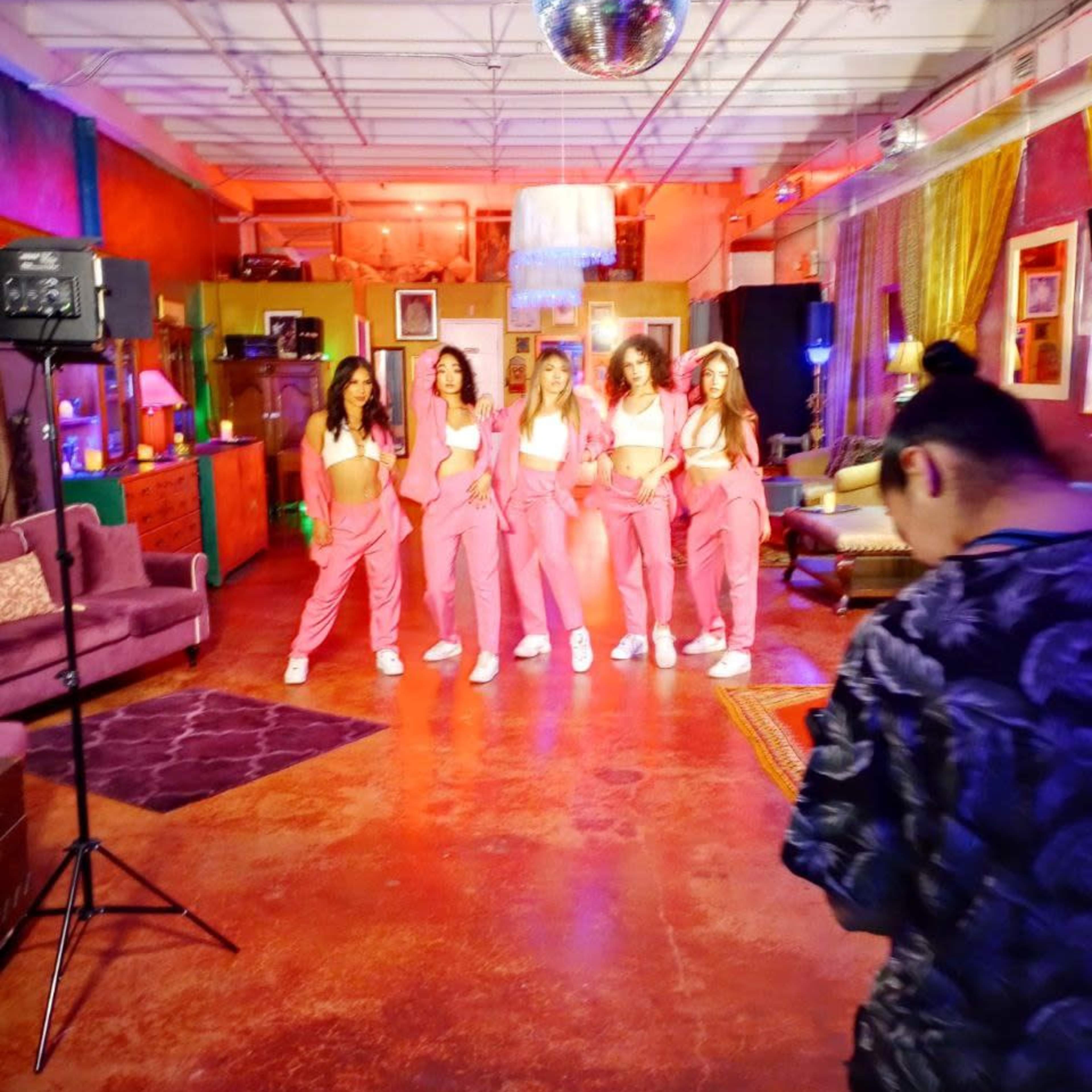A group of five women in matching pink outfits stands in a brightly lit room with colorful decor, while a person in the foreground appears to be taking photos.