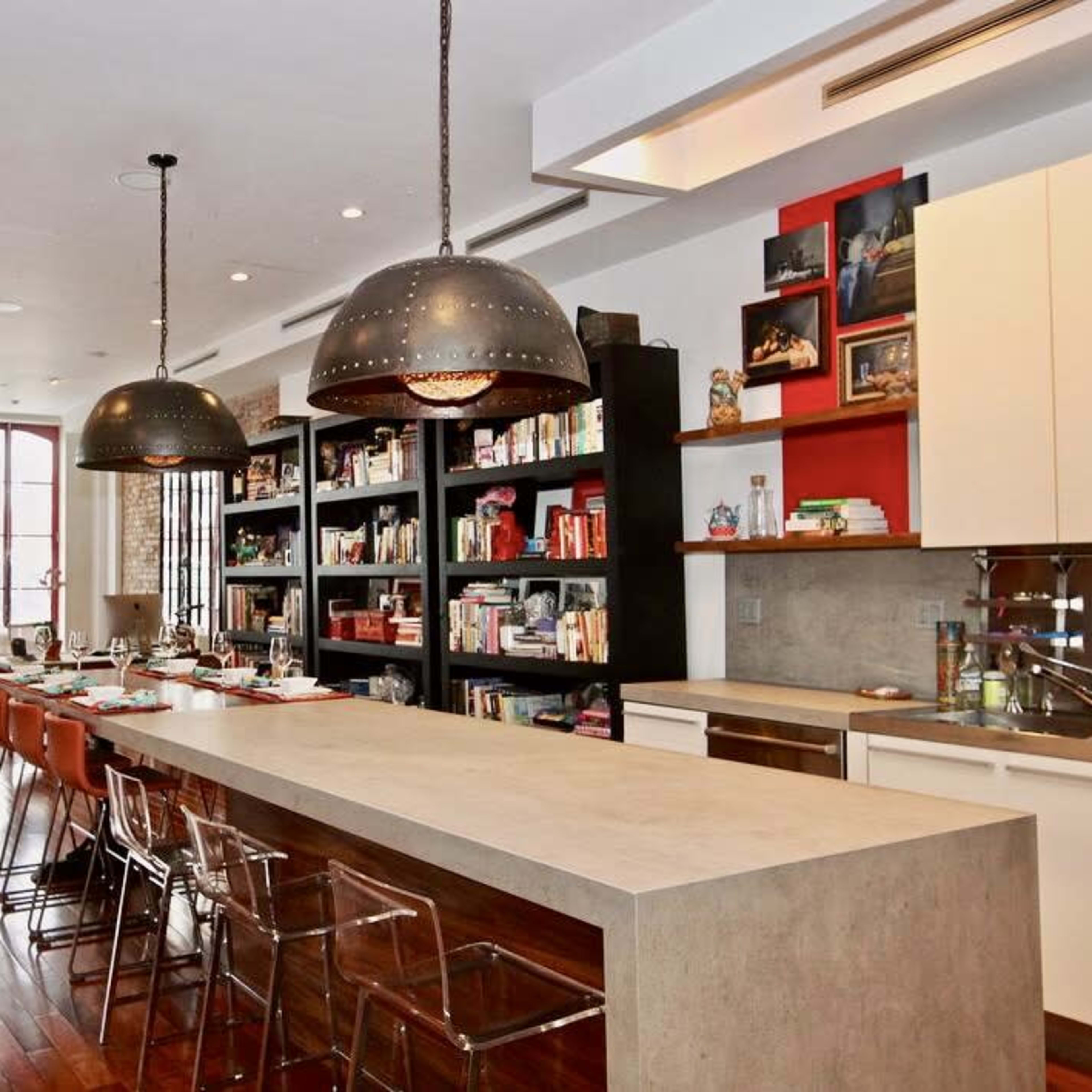 The modern kitchen features a long concrete island with bar seating, surrounded by dark wood cabinets and shelves filled with books and decorative items.