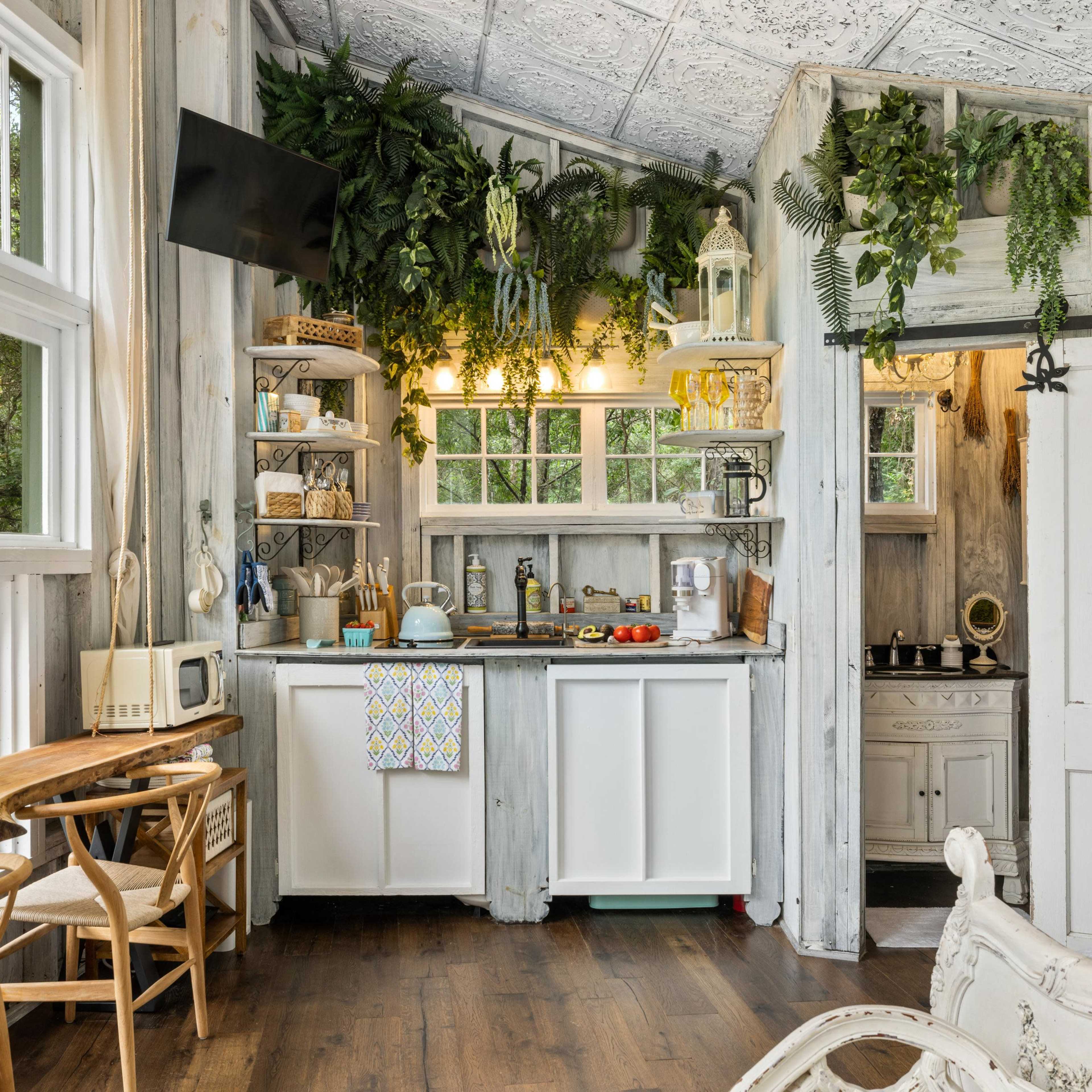 A rustic kitchen with wooden shelves adorned with plants, a countertop with various kitchen items, and a small dining area with light-colored chairs.