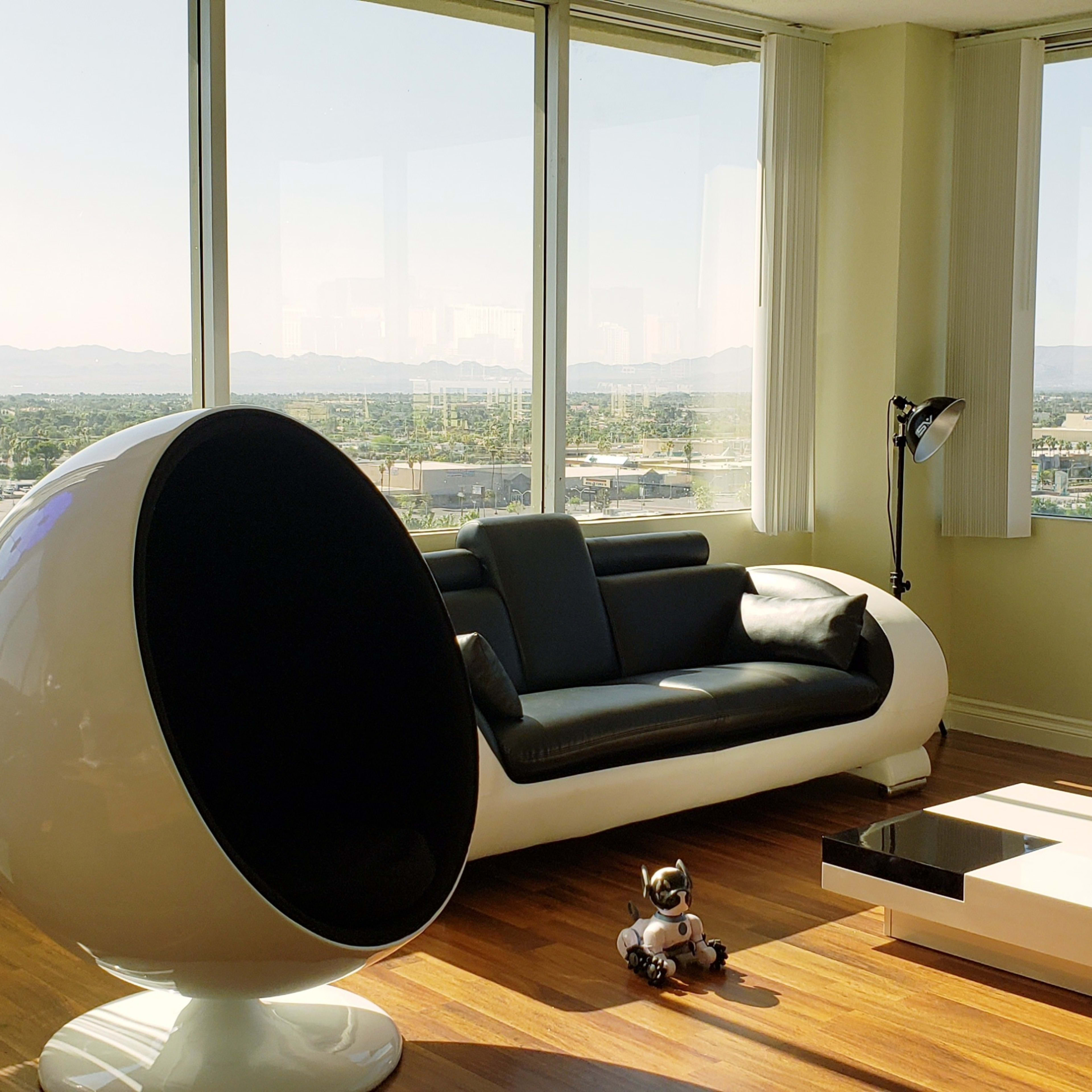 The scene features a modern living room with large windows offering a view of mountains, a black and white sofa, a spherical chair, a coffee table, and a small decorative robot on the wooden floor.