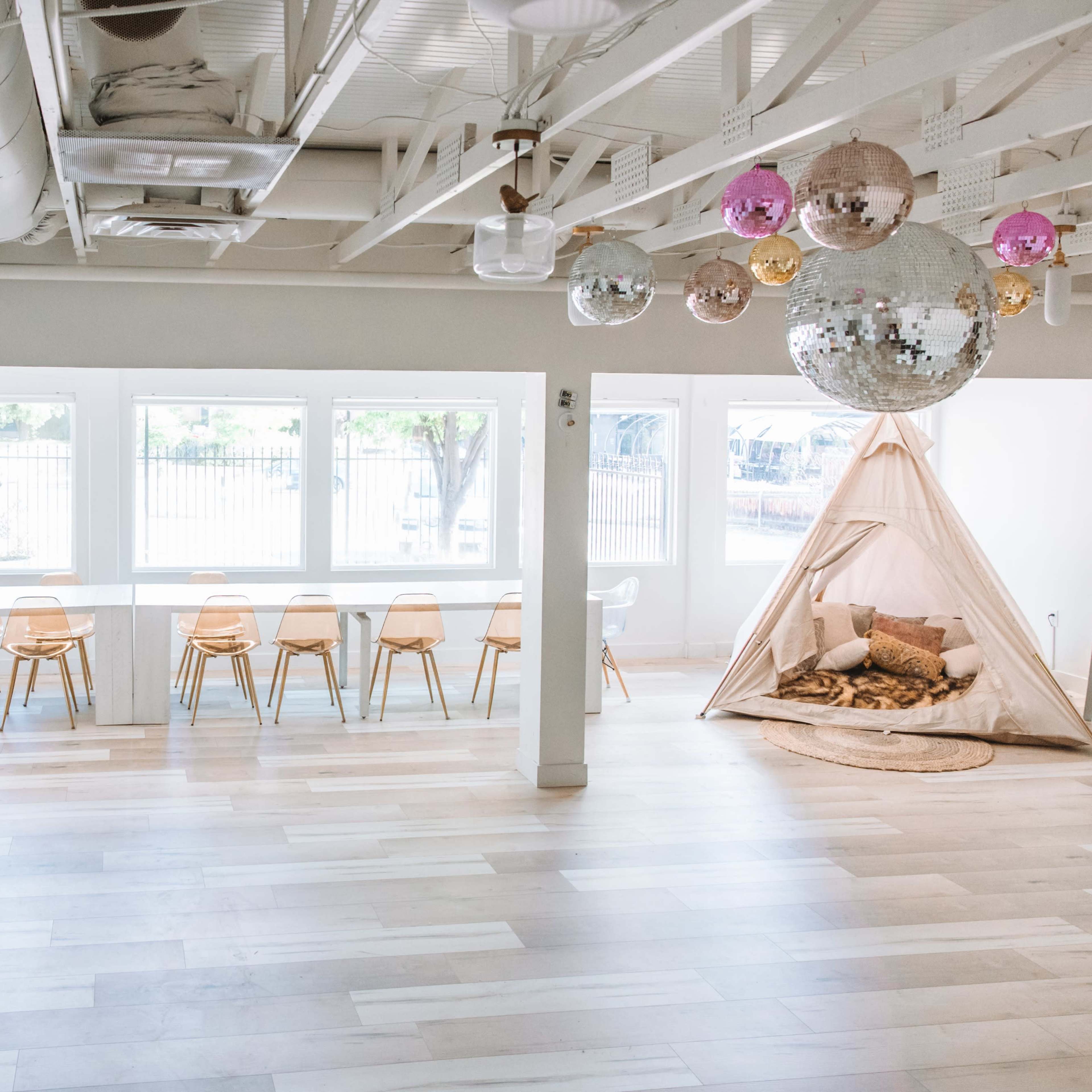 A modern, airy interior space with white walls, light wooden flooring, a long table with chairs, a seating area, and a decorative teepee.