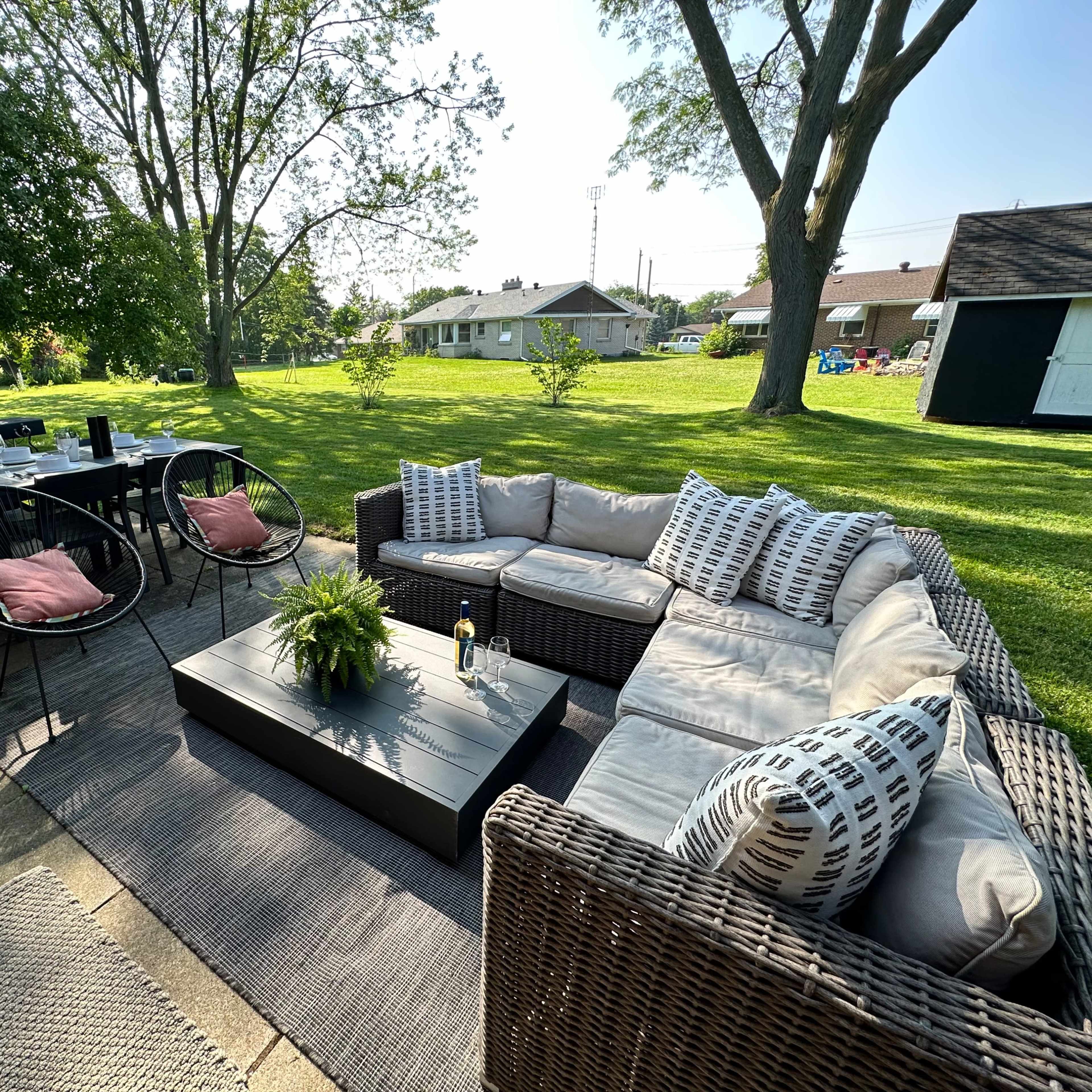 A patio area features a sectional sofa, a coffee table, and a dining table with chairs, set on a grassy lawn surrounded by trees and nearby houses.
