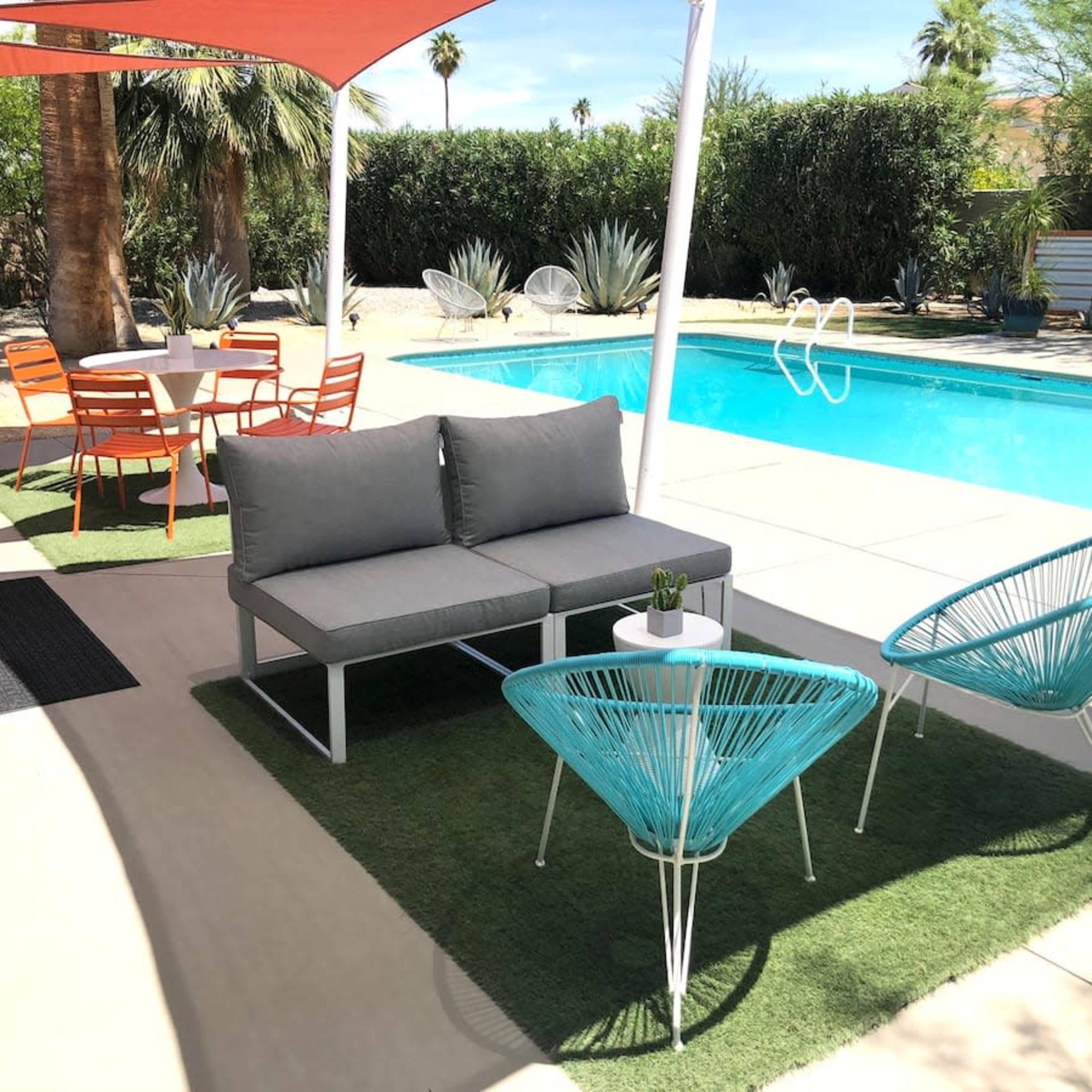 A patio area features a gray sectional sofa and two teal chairs arranged near a swimming pool, with a dining table and chairs situated under a shaded canopy.
