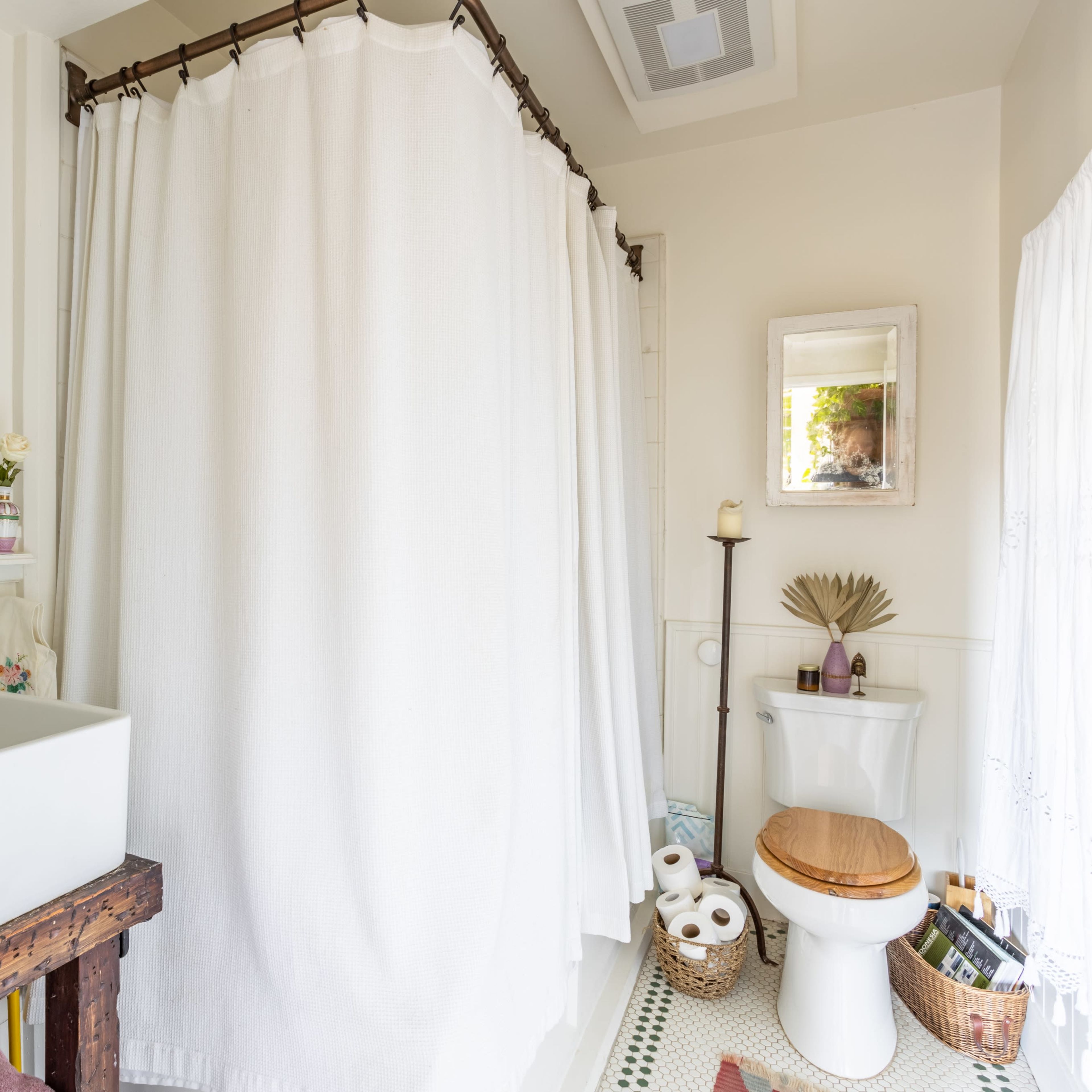 A bright bathroom features a white shower curtain, a sink on a wooden stand, a toilet with a wooden seat, and decorative elements like towels and toiletries.