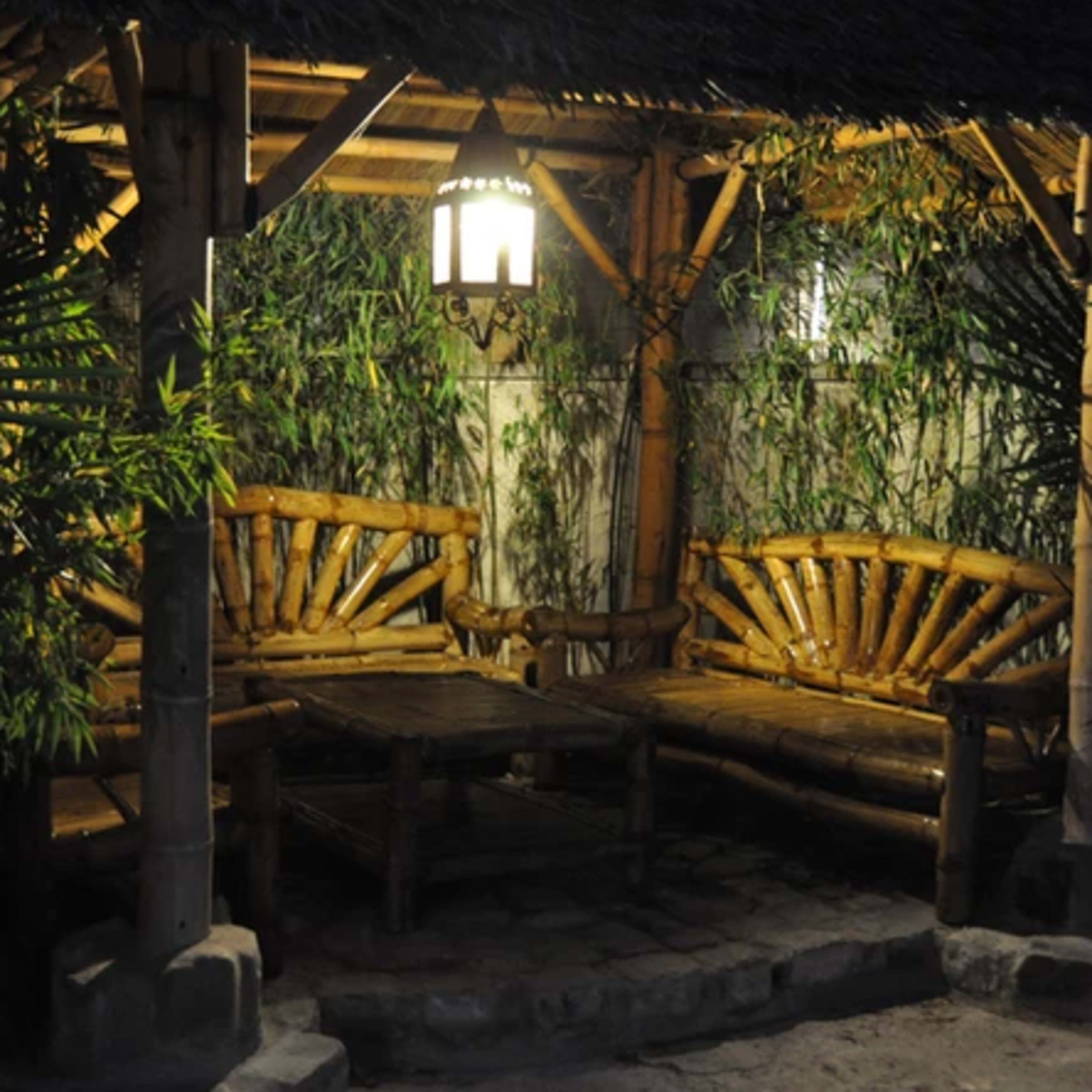 A bamboo patio with two curved benches and a table, surrounded by greenery and illuminated by a hanging lantern.