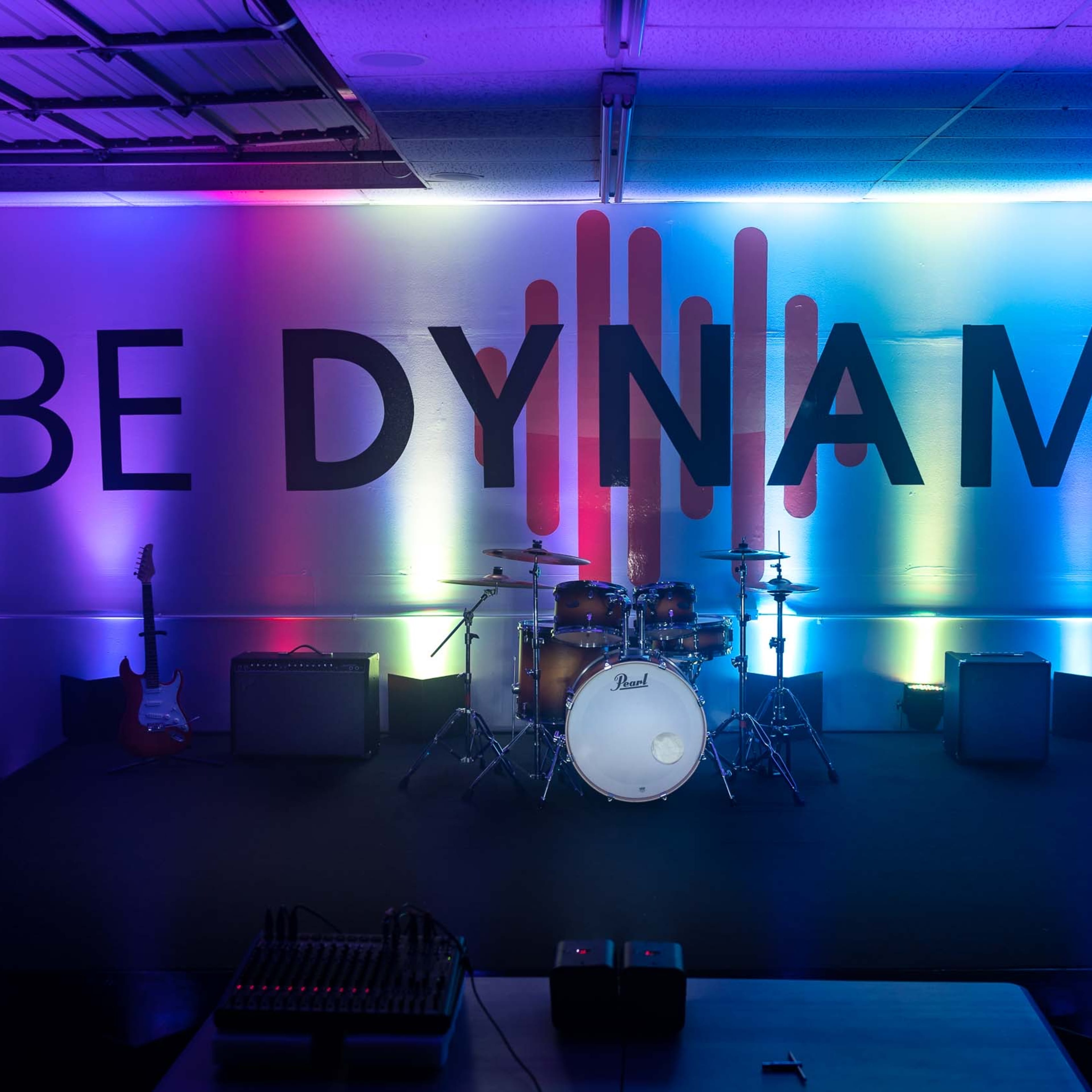 A stage setup with musical instruments in front of a brightly lit backdrop featuring the phrase "BE DYNAMIC" in large letters.