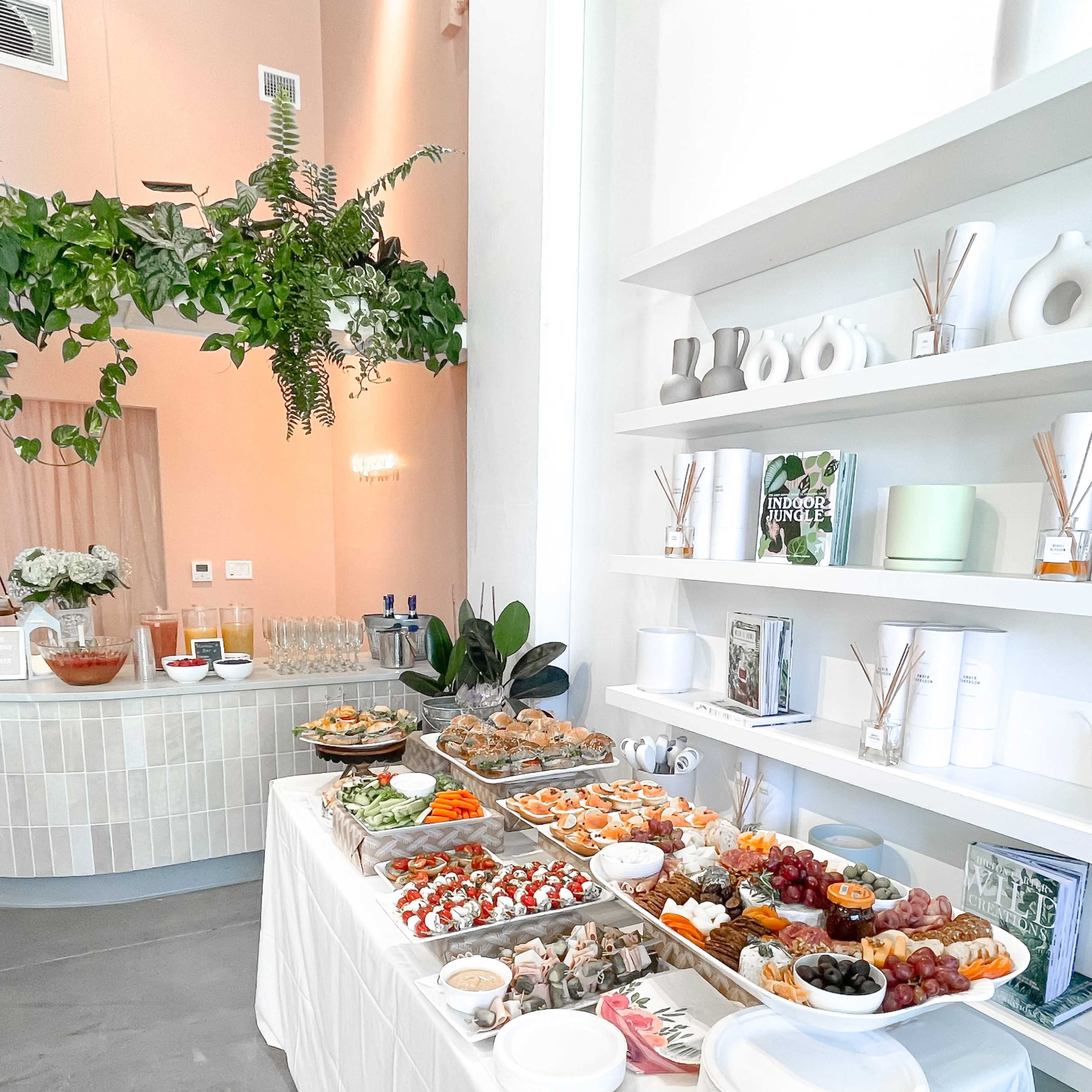 A white tablecloth displays a variety of food platters, including seafood, fruits, and pastries, against a backdrop of decorative shelves and greenery.