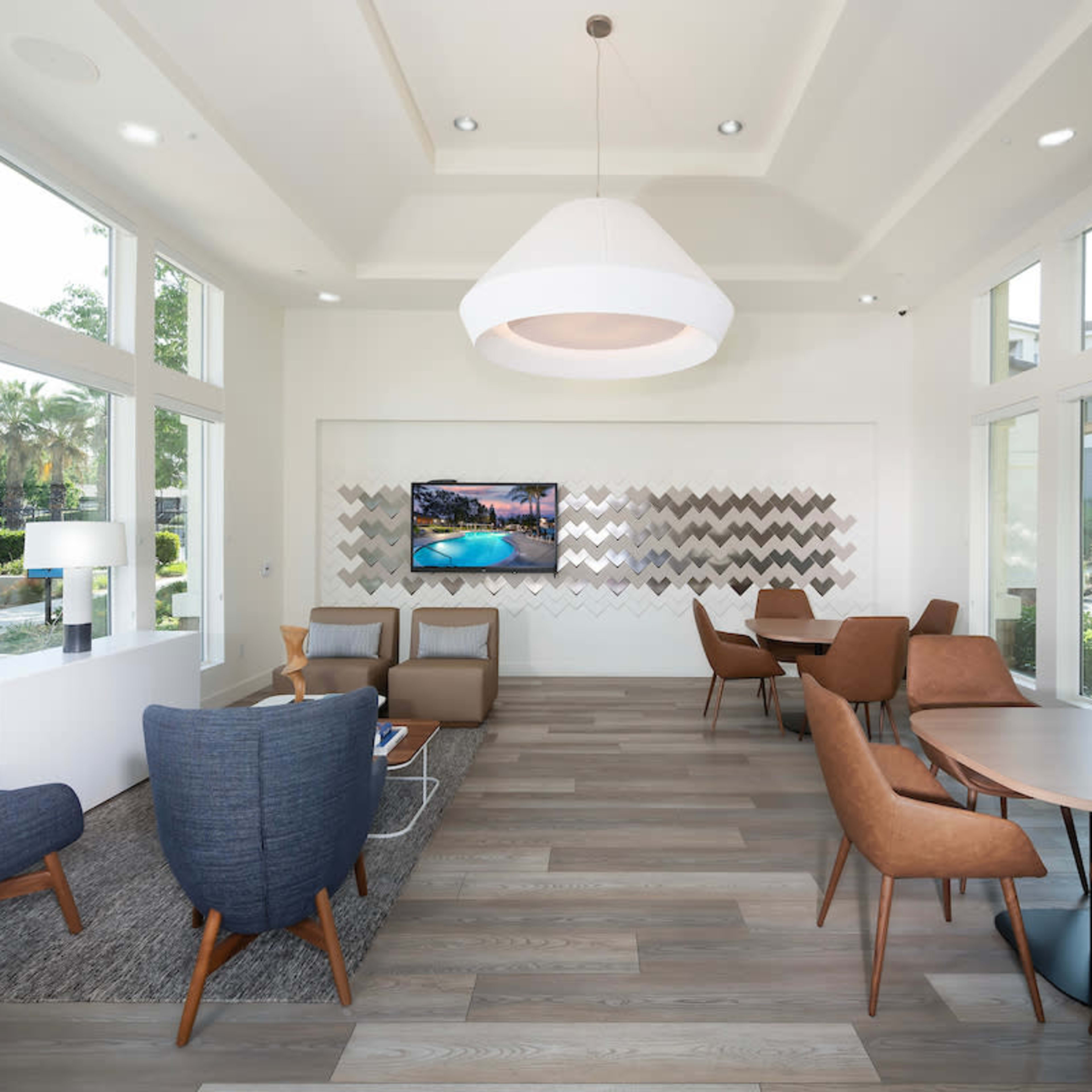 A modern lounge area featuring a mix of armchairs and dining tables, large windows, and a wall-mounted television.