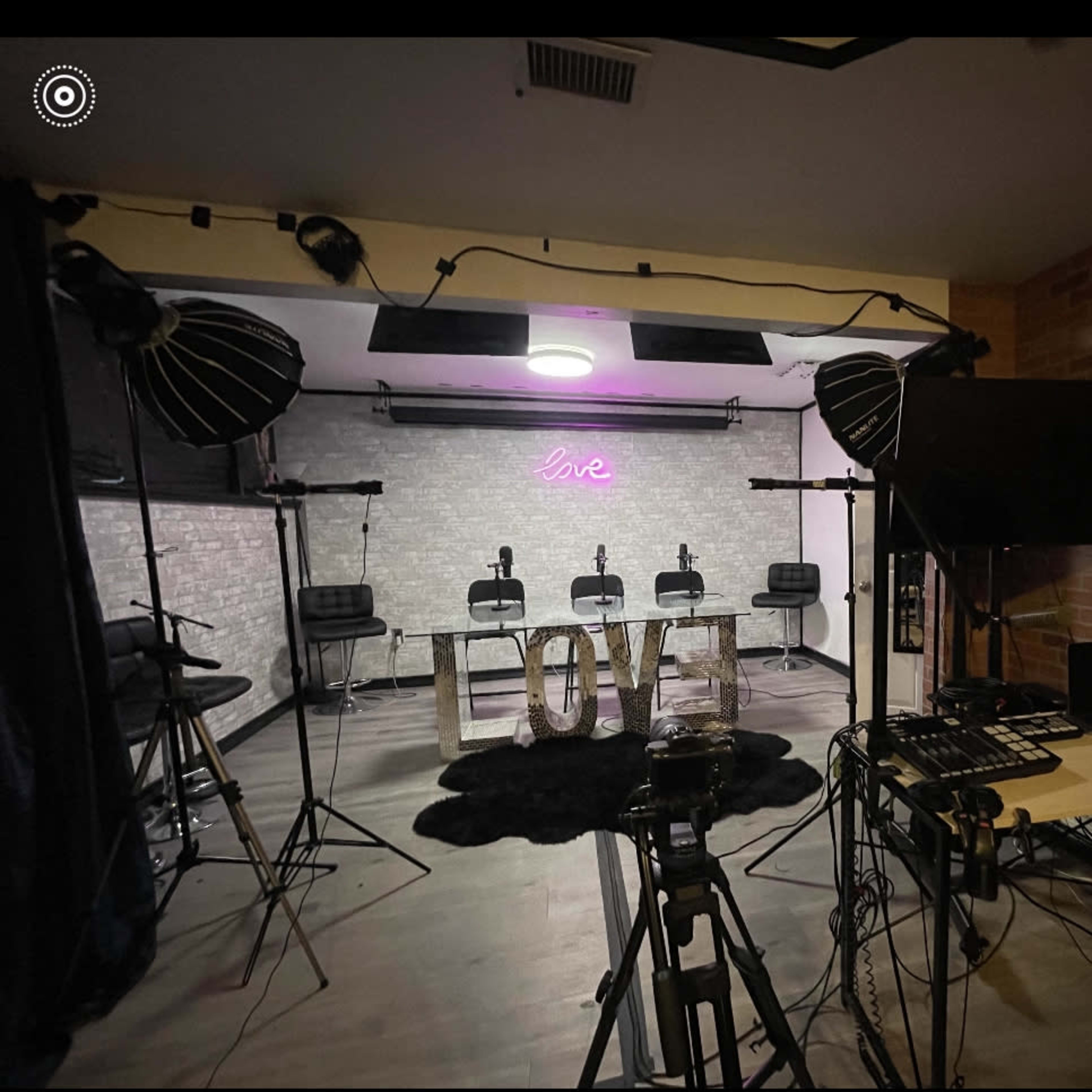 A studio setup with four chairs facing a lit "live" sign, surrounded by professional lighting equipment and a sound mixer.