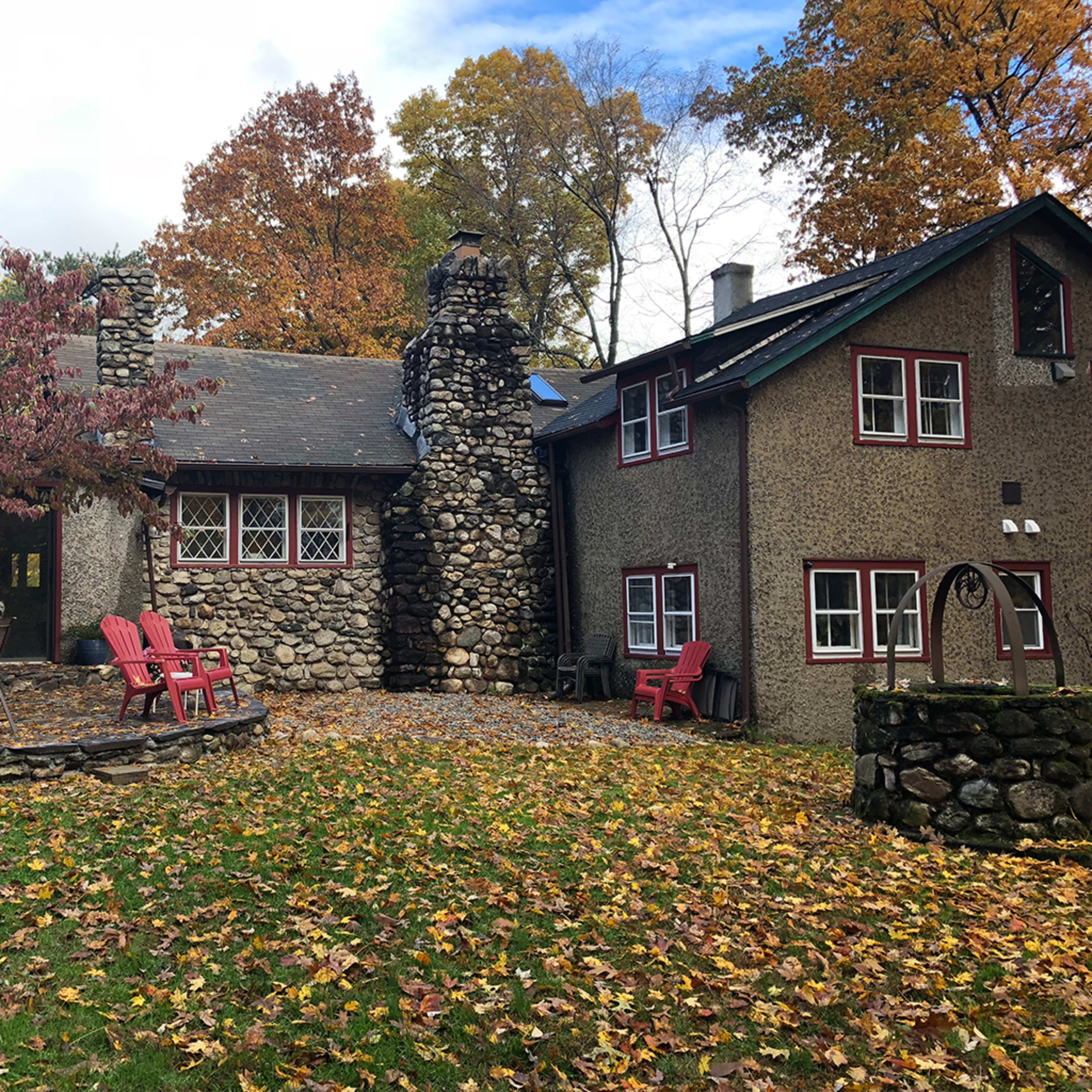 A stone cottage with a large chimney, surrounded by trees and a yard covered in fallen leaves, features outdoor seating in a cozy arrangement.