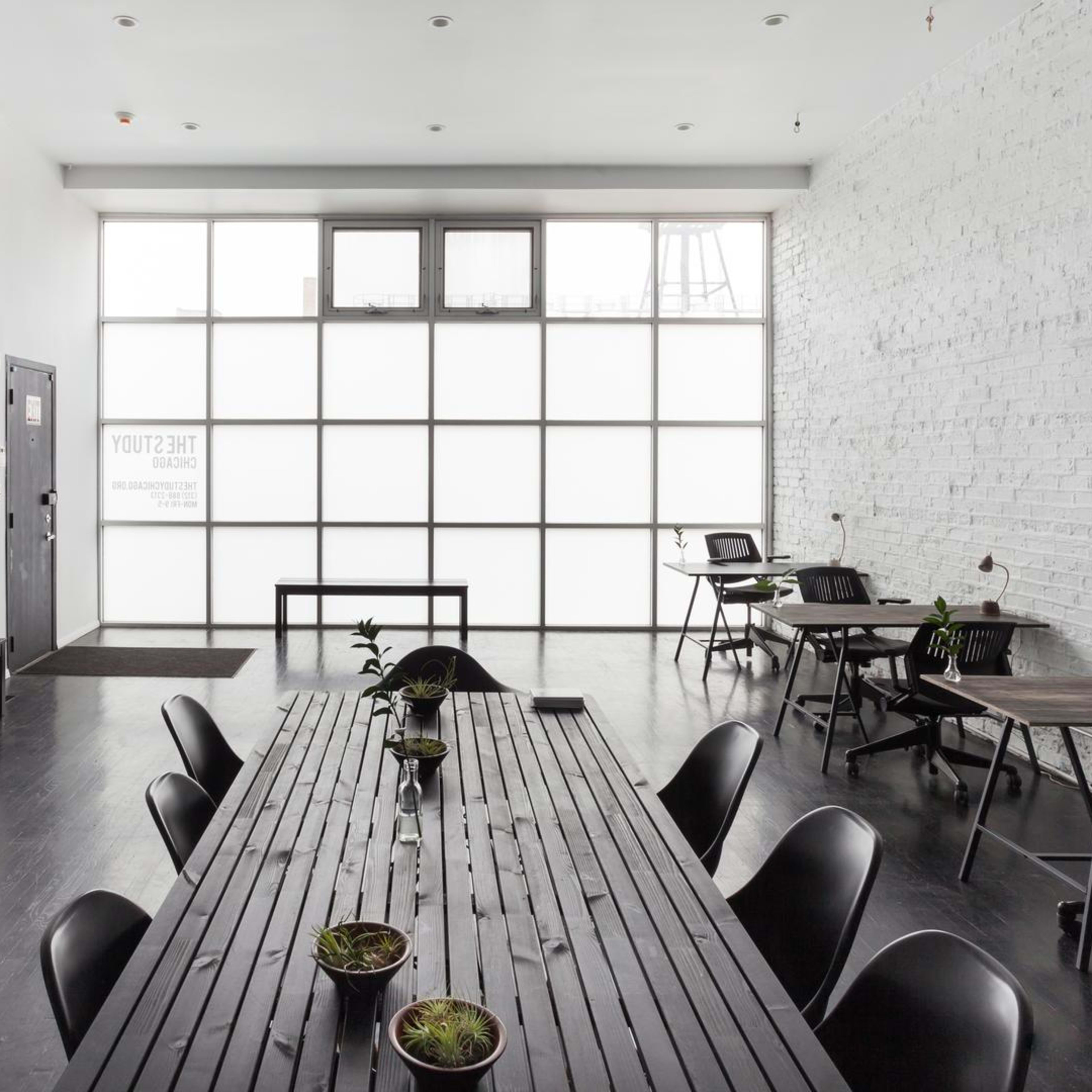 A minimalist meeting room featuring a long wooden table, black chairs, and several small plants, with large windows letting in natural light.