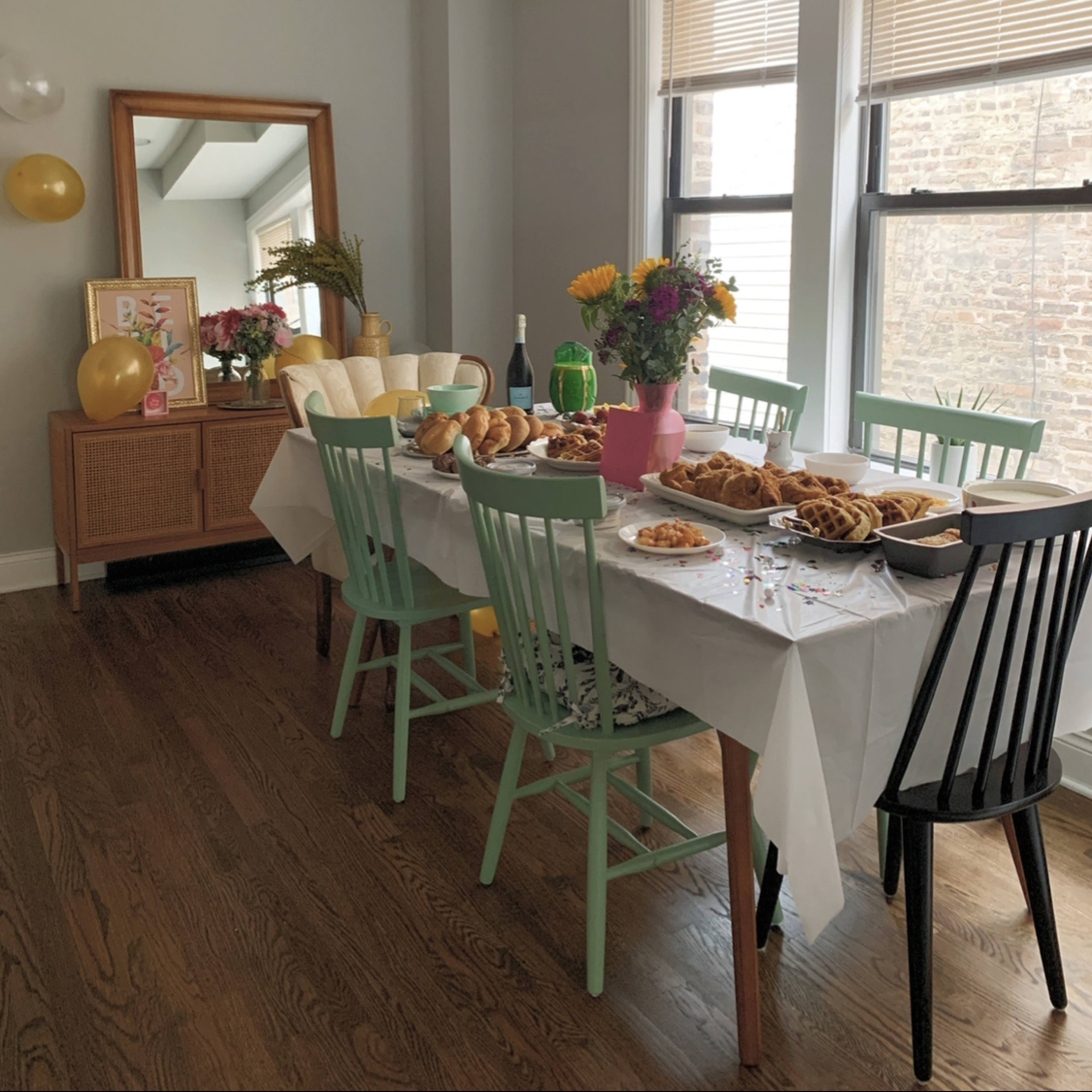 A dining table is set with an assortment of food and drinks, surrounded by chairs in a brightly lit room with large windows.