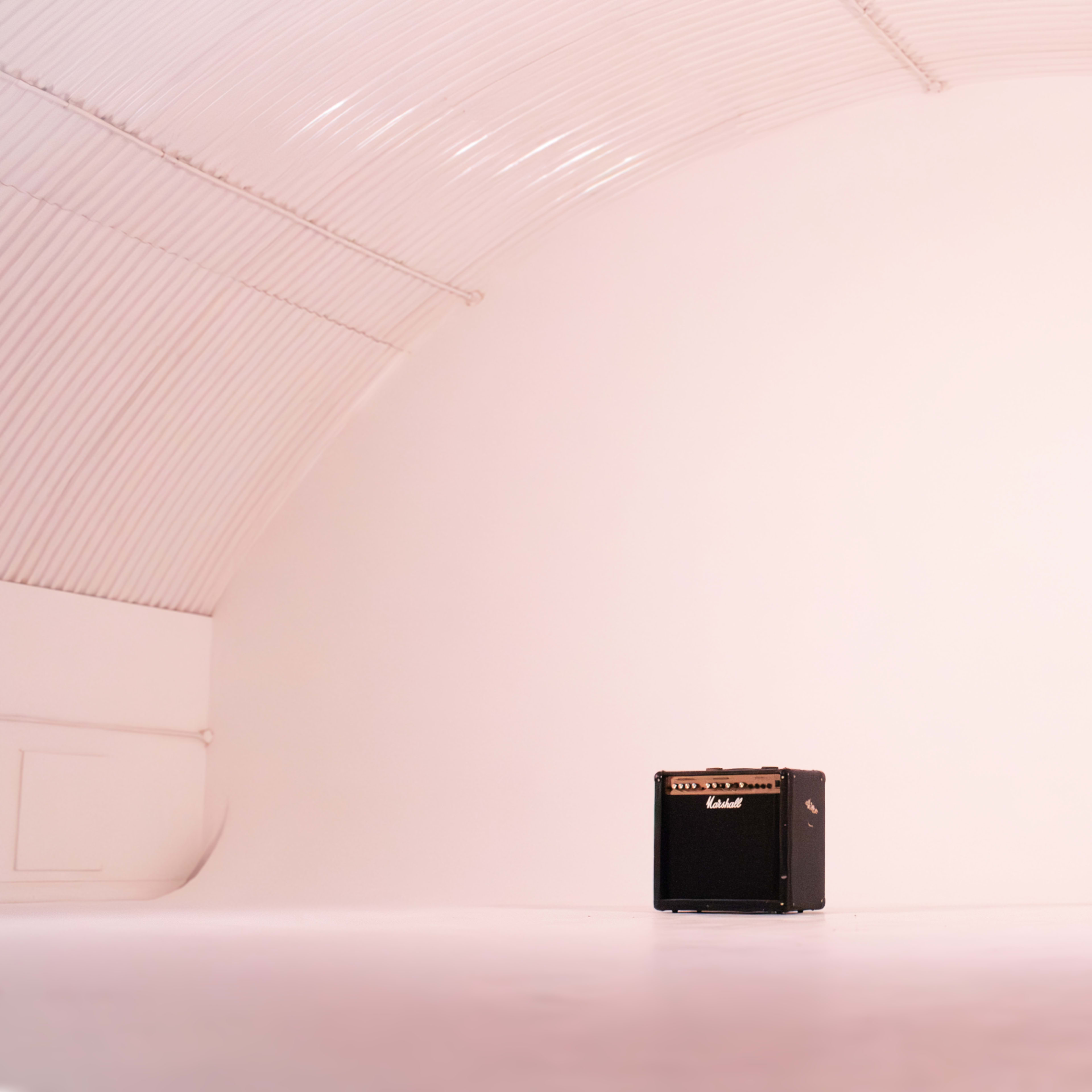 A black guitar amplifier stands alone on a blank white floor within a curved white-walled space.