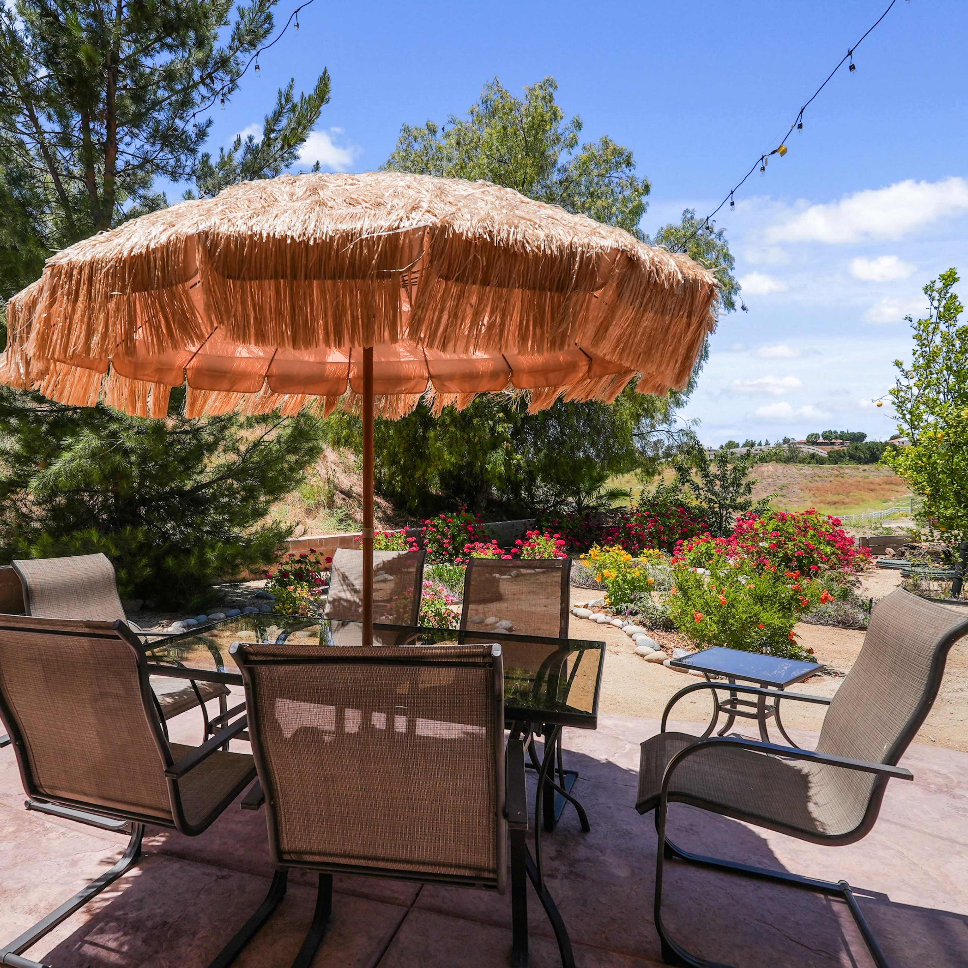 A shaded patio area features a thatched umbrella over a table with surrounding chairs, set against a backdrop of gardens and trees.