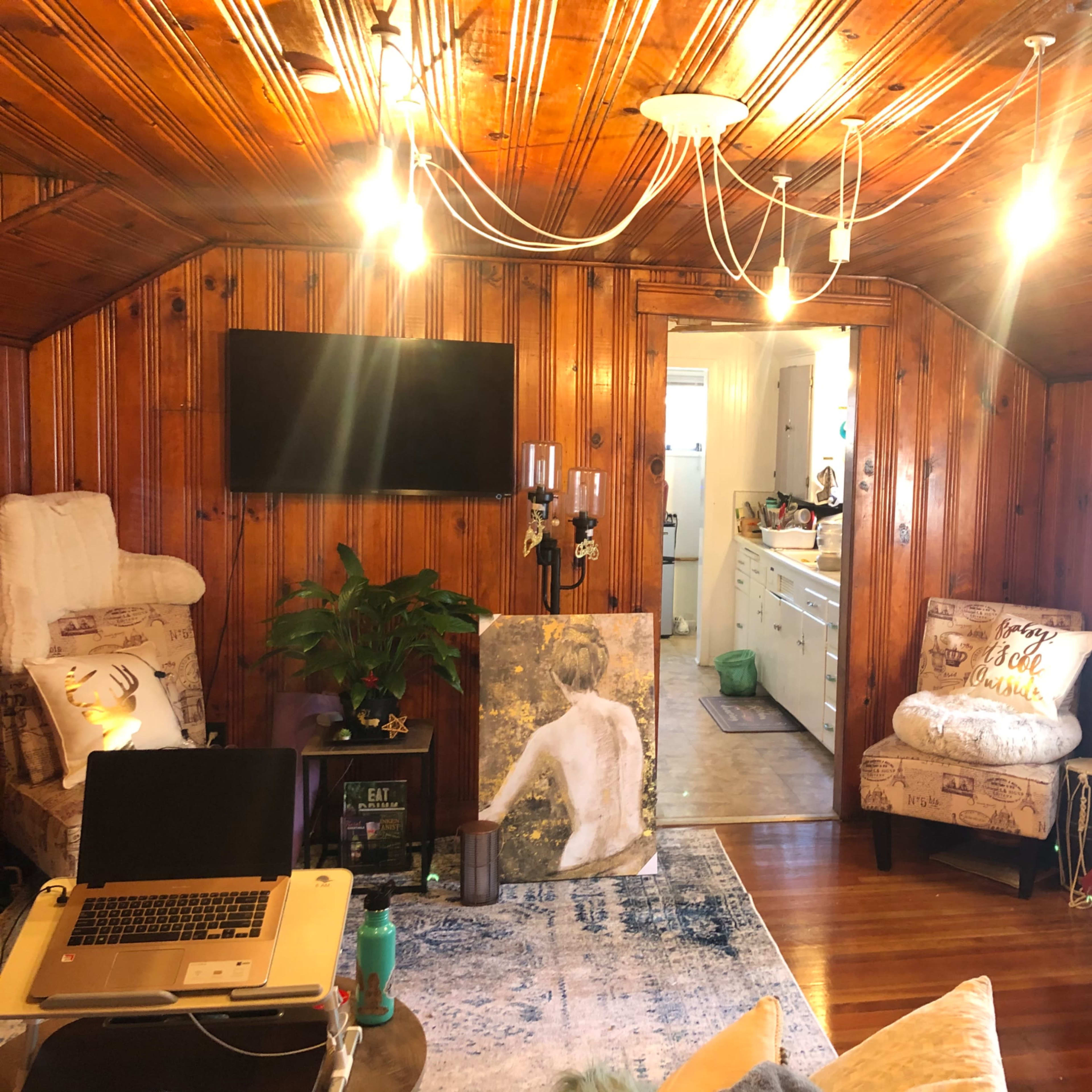 The room features wooden paneled walls, a small seating area with a laptop, a large decorative painting, and an entryway leading to a kitchen visible in the background.