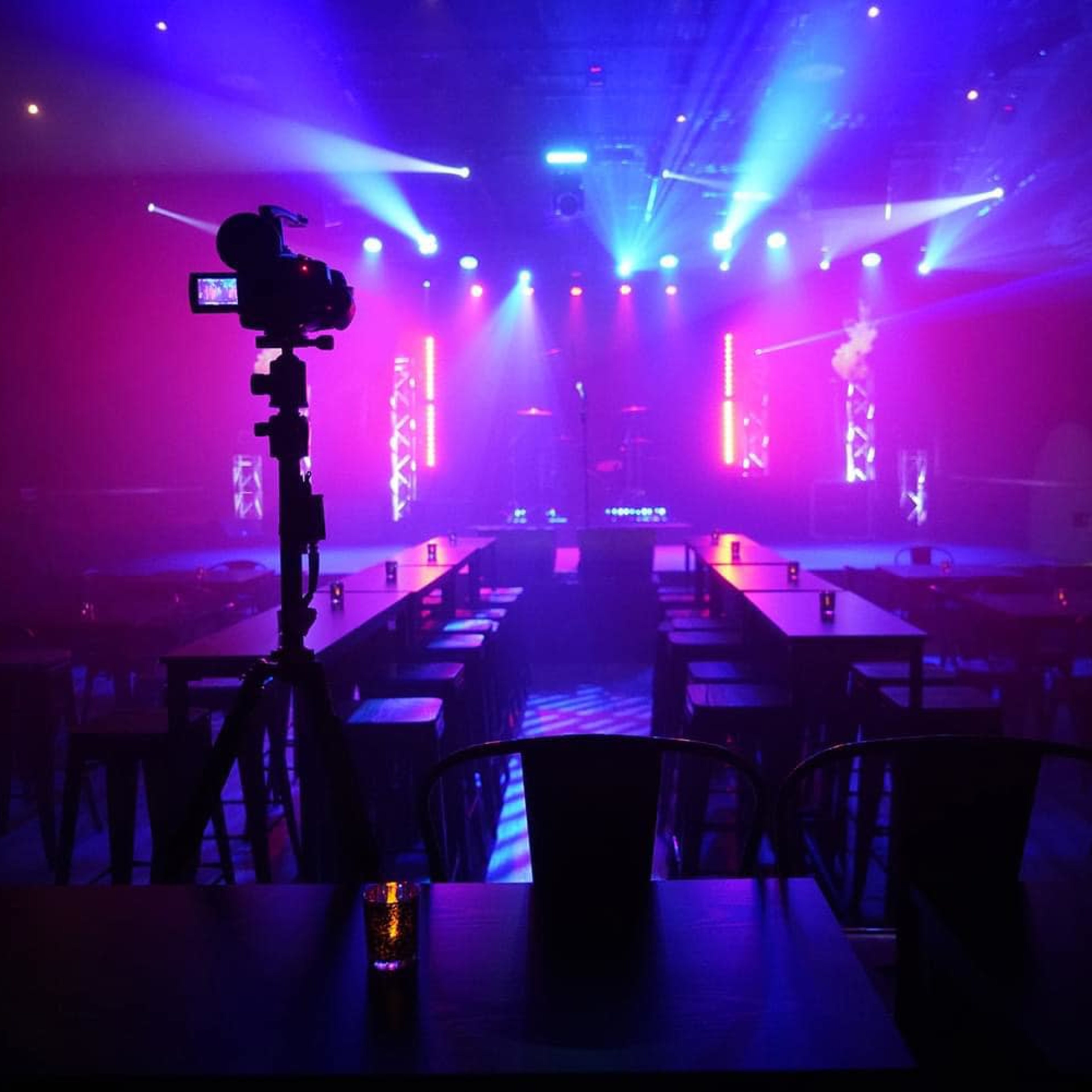 A stage is set with colorful lights and a camera positioned in front, while tables are arranged in rows with candles placed on them.