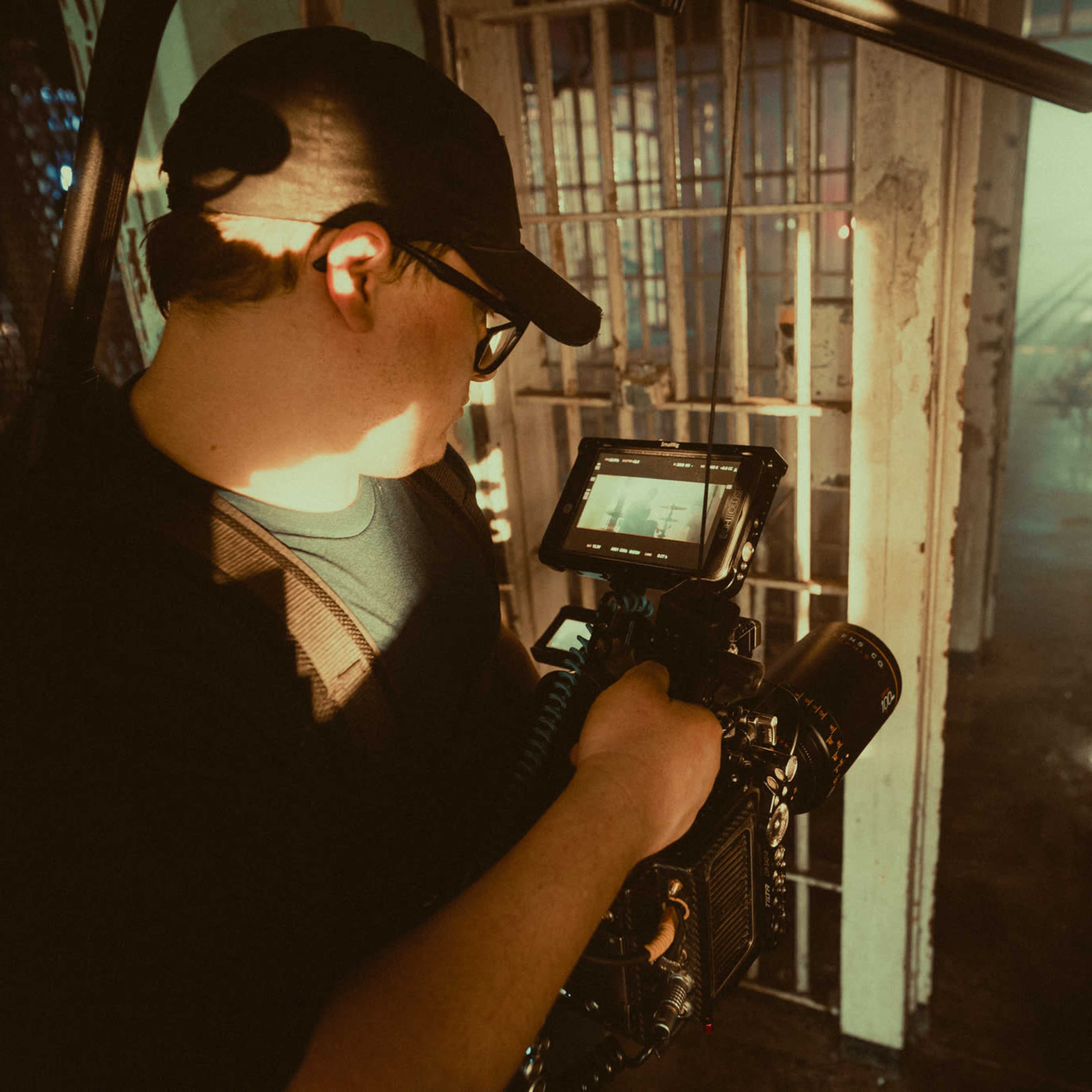 A person with glasses and a black cap operates a camera in a dimly lit space resembling a prison.