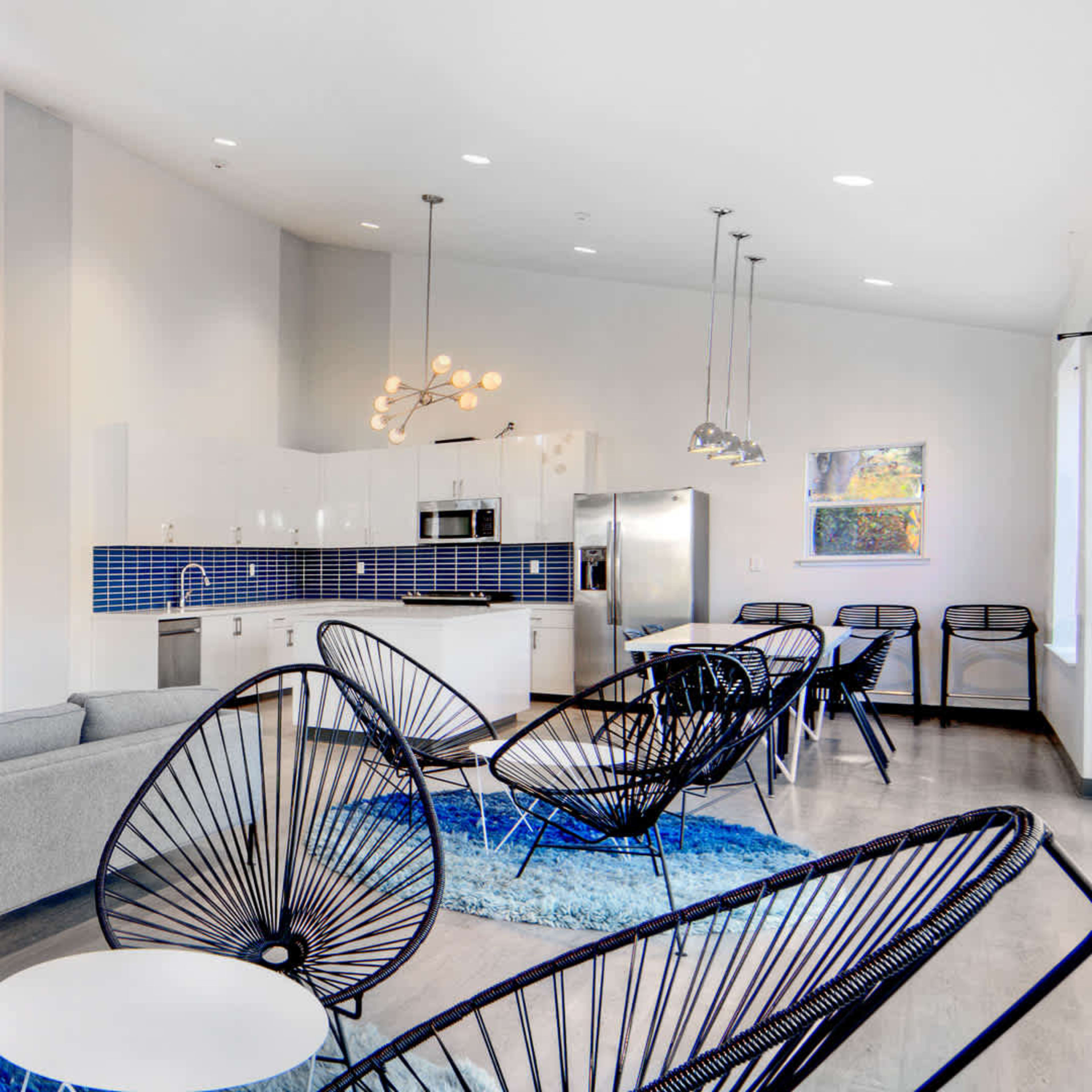 A bright and modern lounge area features a gray sofa, black chairs, a blue rug, and a kitchen with a blue backsplash and stainless-steel appliances.